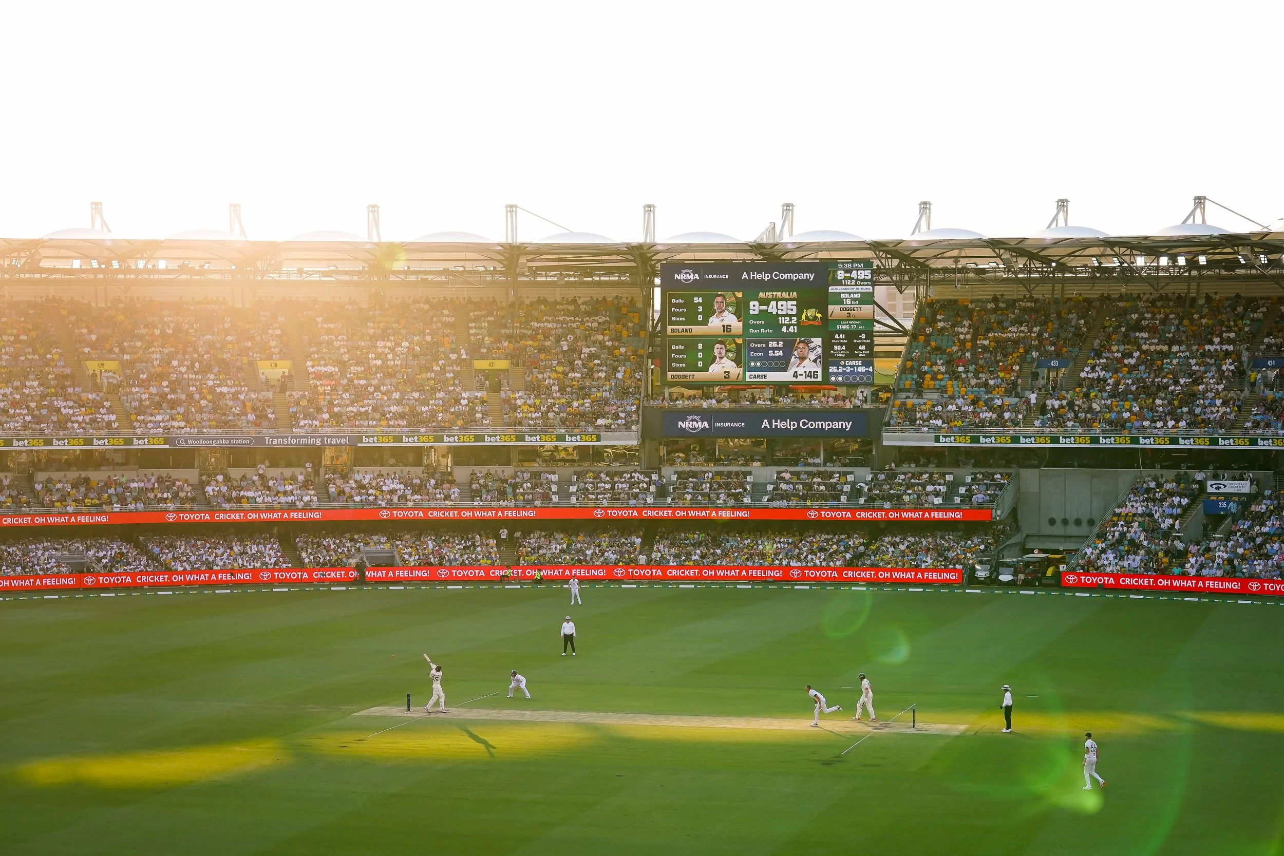 Cricket players on the field in a stadium, with a large crowd watching and a scoreboard displaying game details.