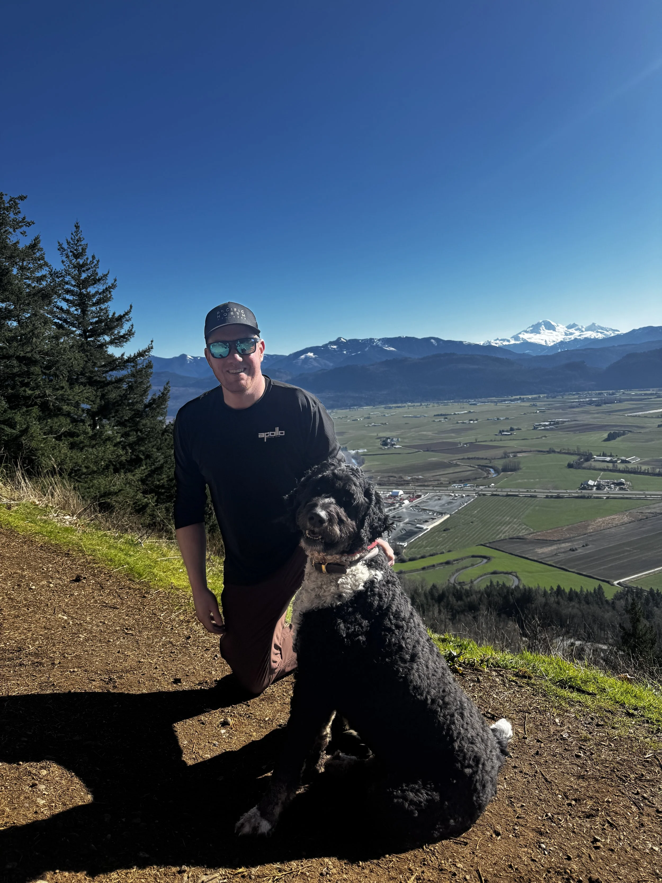 A man in sunglasses and a cap smiling while kneeling on a dirt trail with a black and white dog in front of him. They are on a hillside overlooking lush fields and mountains with snow-capped peaks in the distance under a clear blue sky.
