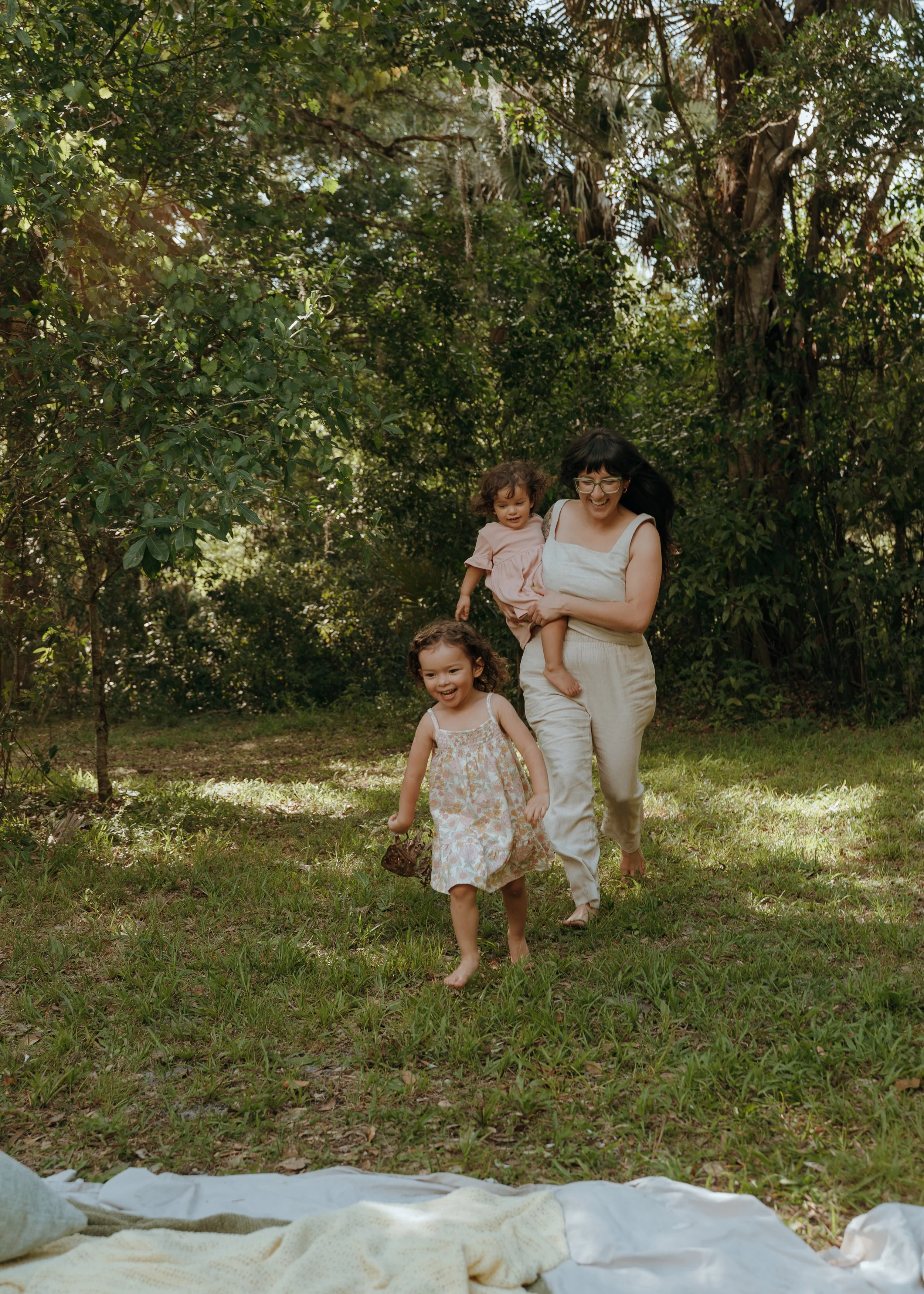 Mother laughing as she follows her two young children through a wooded park during a candid Palm Beach family session.