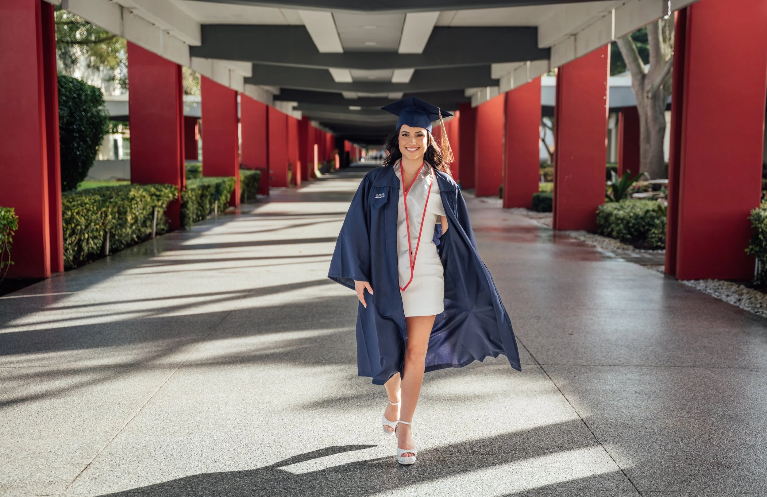 FAU graduate smiling as she walks through the red-columned campus walkway in Boca Raton, Florida, wearing her navy blue cap and gown.