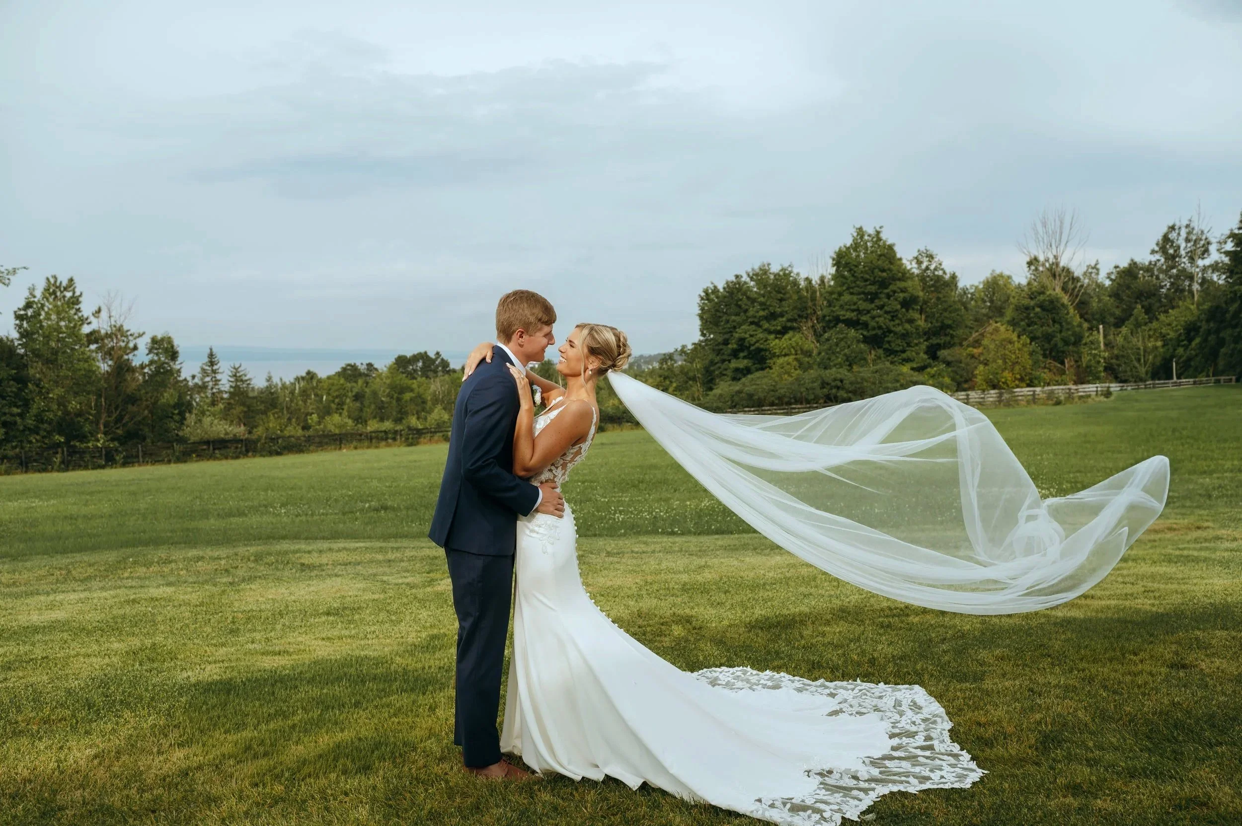 Sydney and Matt kiss in a field as her veil blows behind them during their Petoskey wedding portraits.