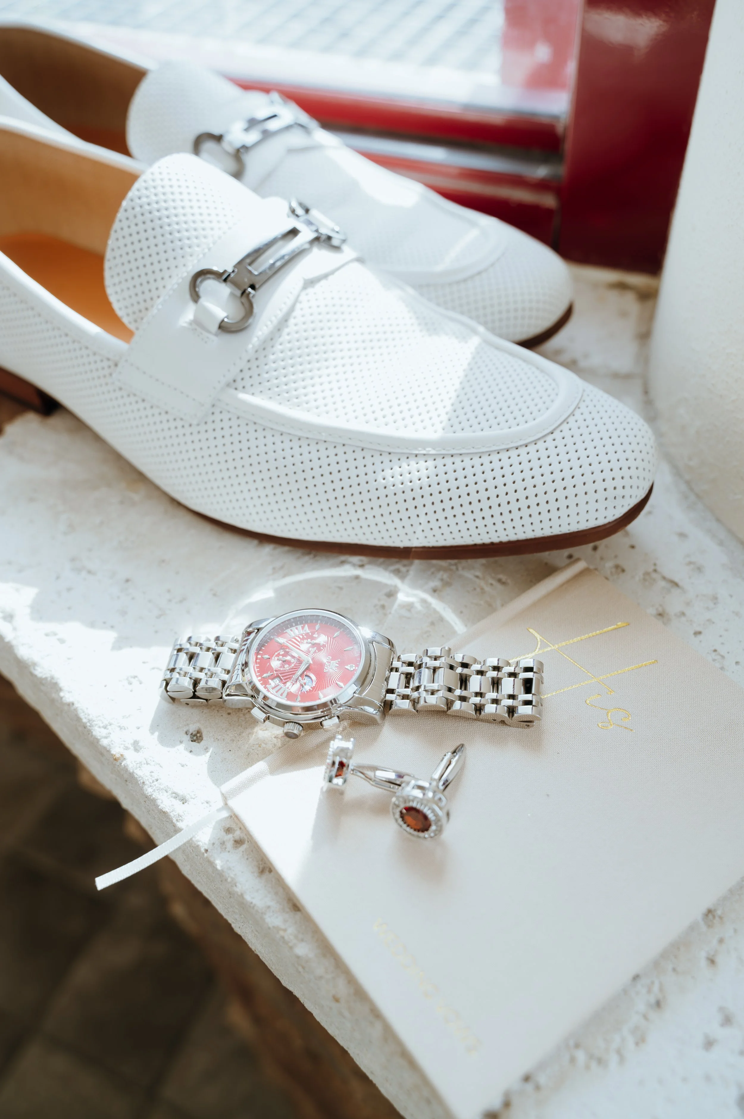 White groom's shoes, silver watch with a burgundy face, and cufflinks arranged with a wedding vow book.