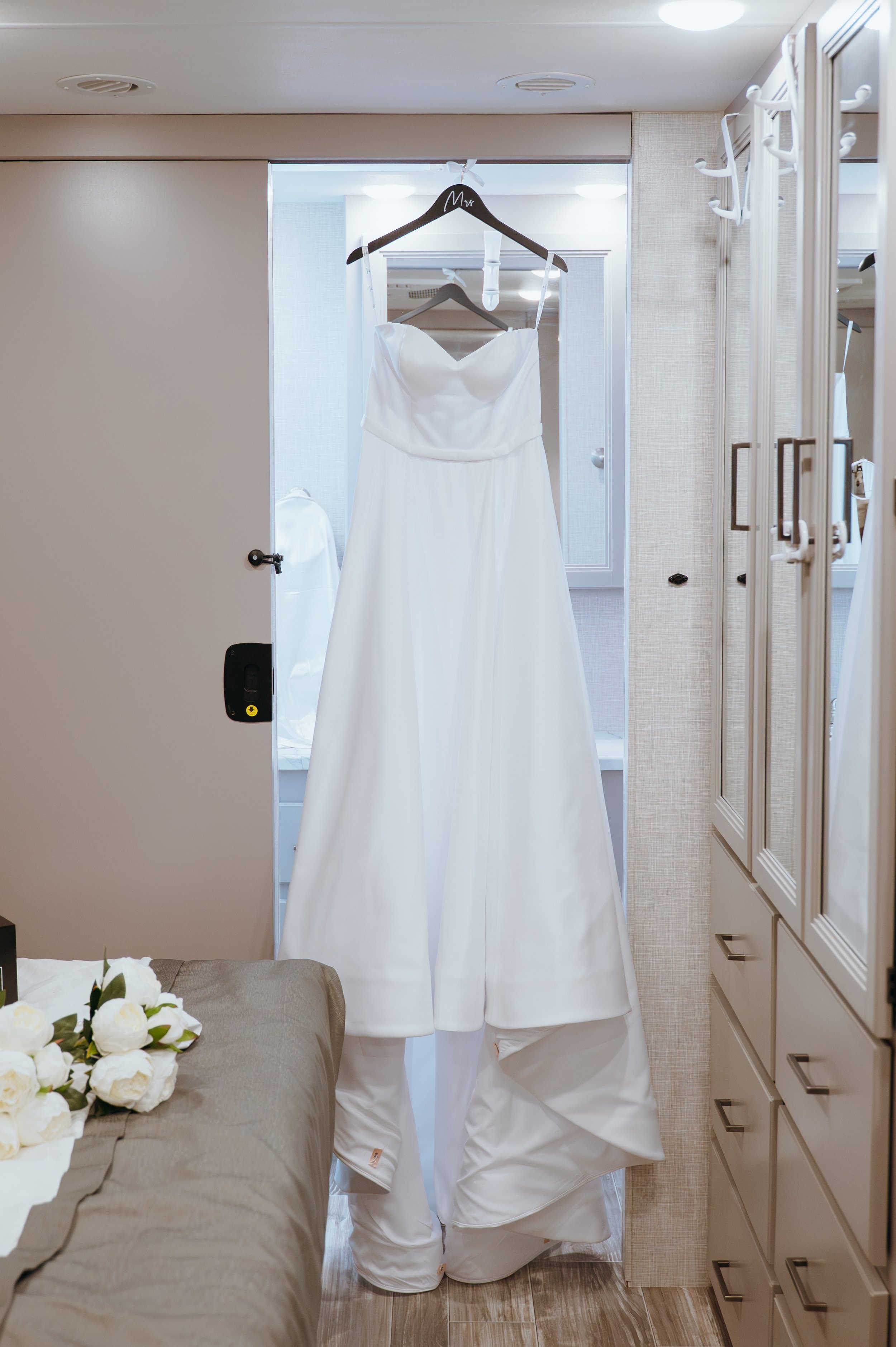 Anndi's white wedding dress hanging beside a bouquet of white roses before her Royal Palm Beach wedding.