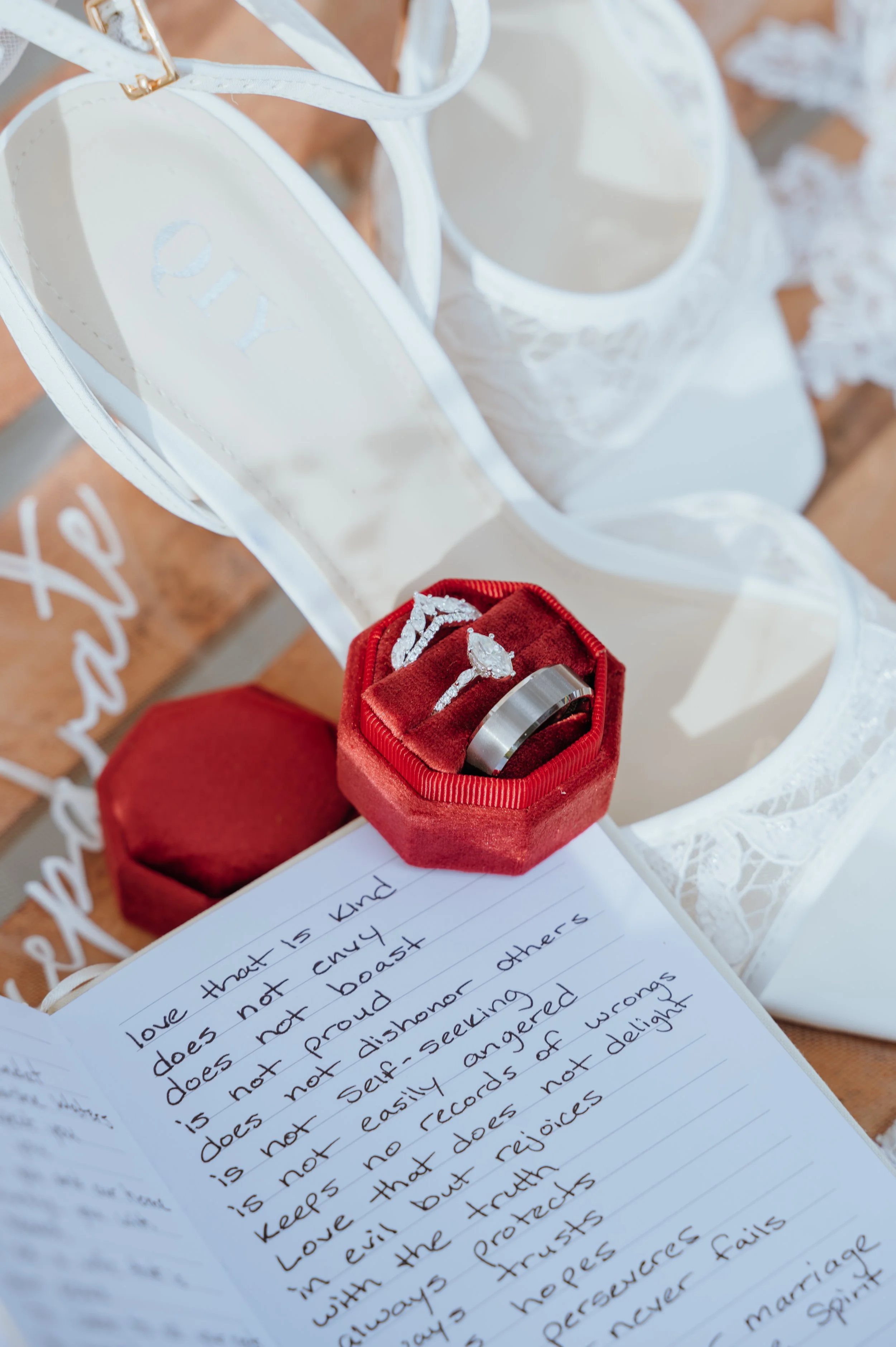 Wedding rings in a burgundy velvet box beside handwritten vows and white bridal heels.
