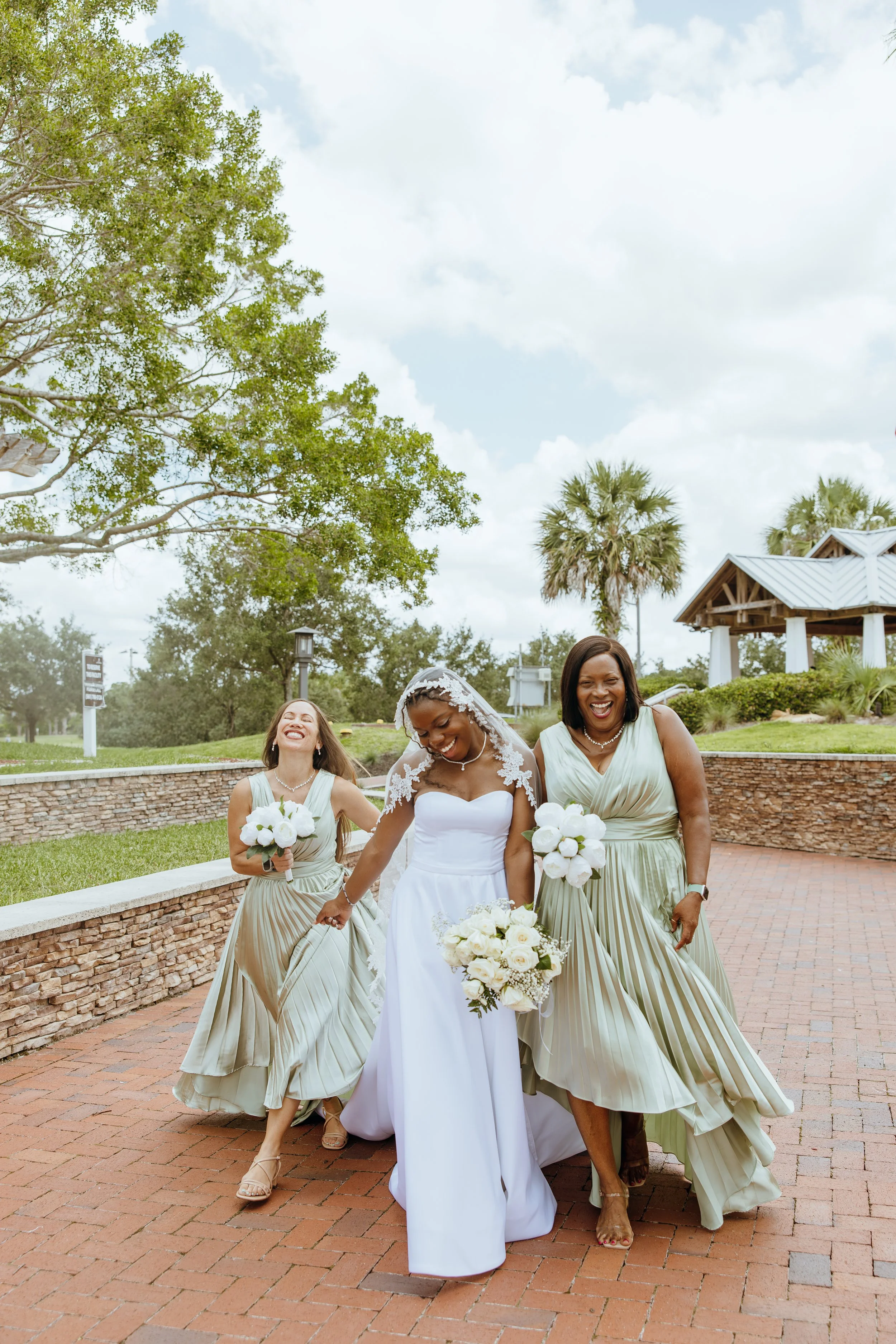 Bride Anndi Waters laughing with two bridesmaids in flowing sage green dresses during wedding party portraits.
