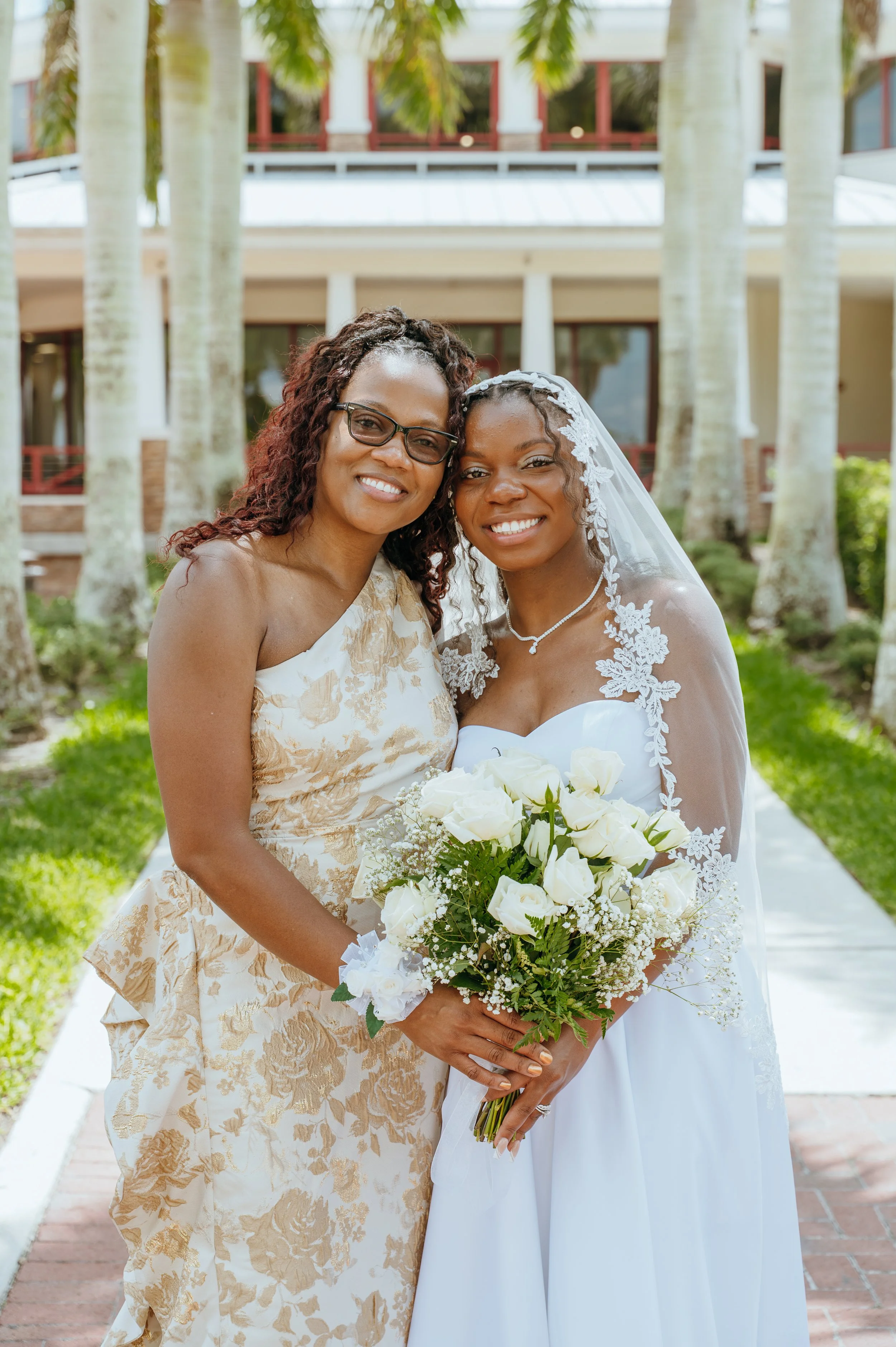 Anndi Waters smiling with a loved one in a cream and gold floral dress while holding white wedding flowers.