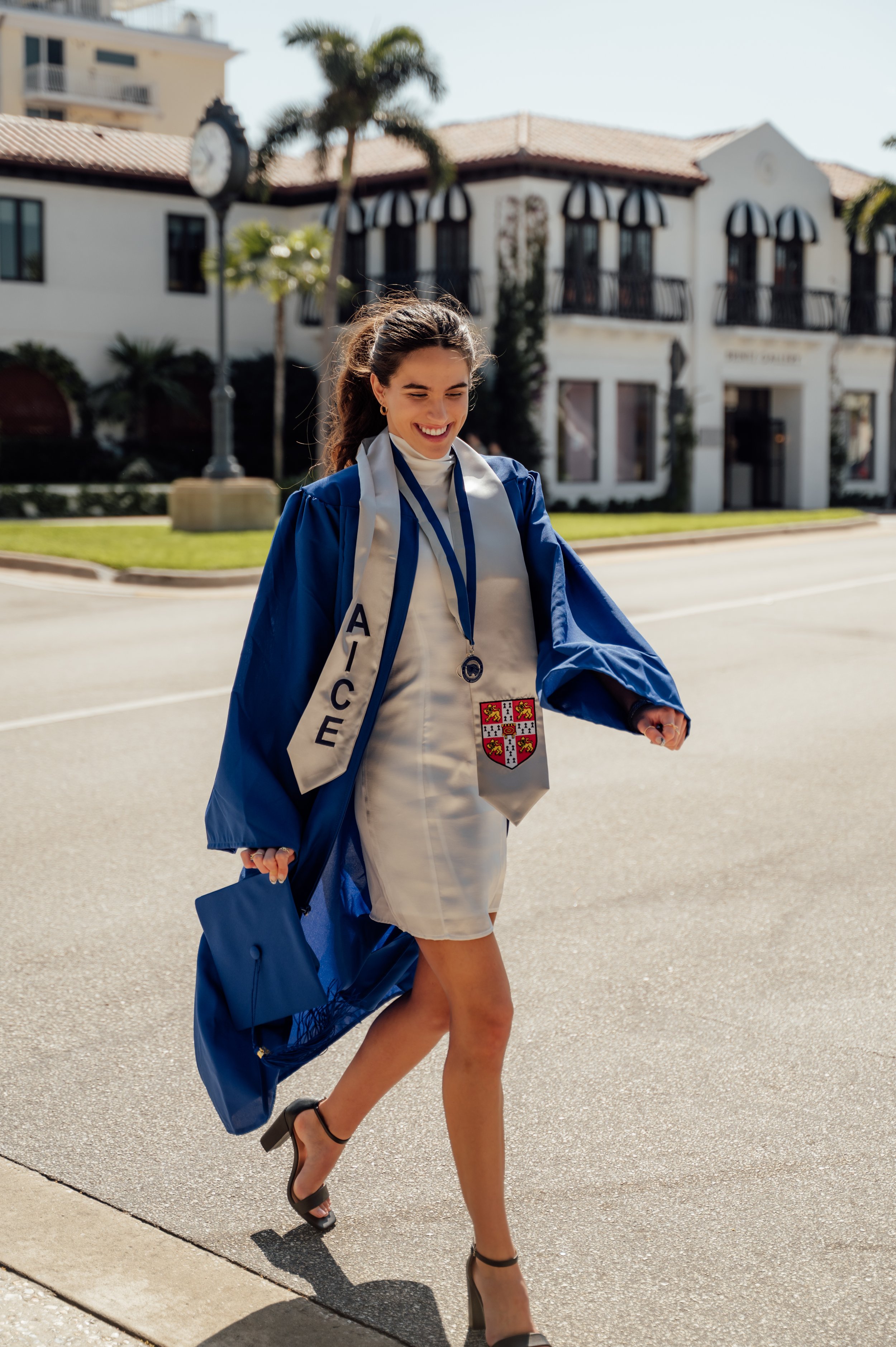 High school senior smiling as she walks along Atlantic Avenue in Delray Beach wearing her graduation gown and stole.