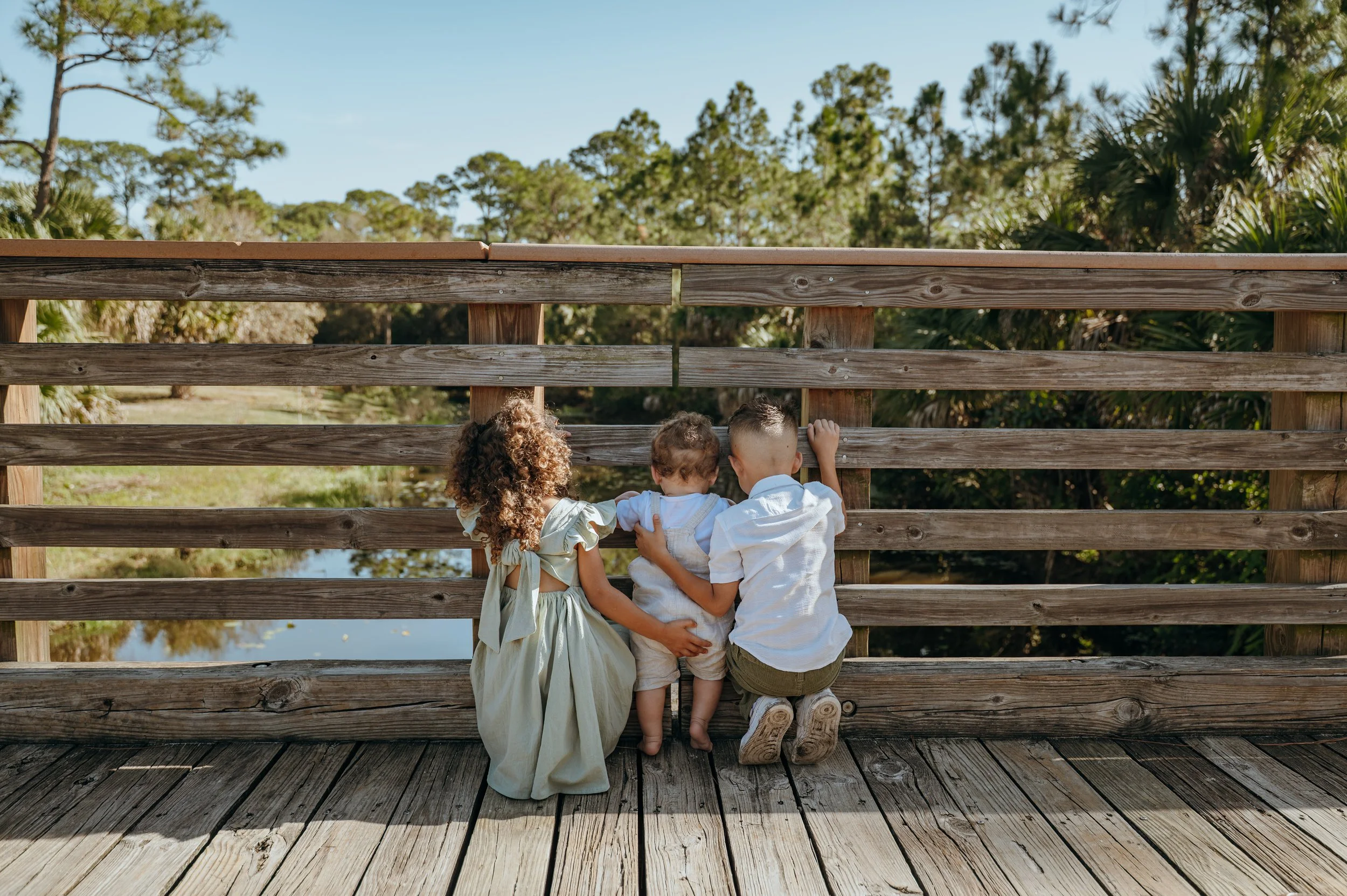 Three siblings standing together at a wooden railing during an outdoor Palm Beach family photography session.