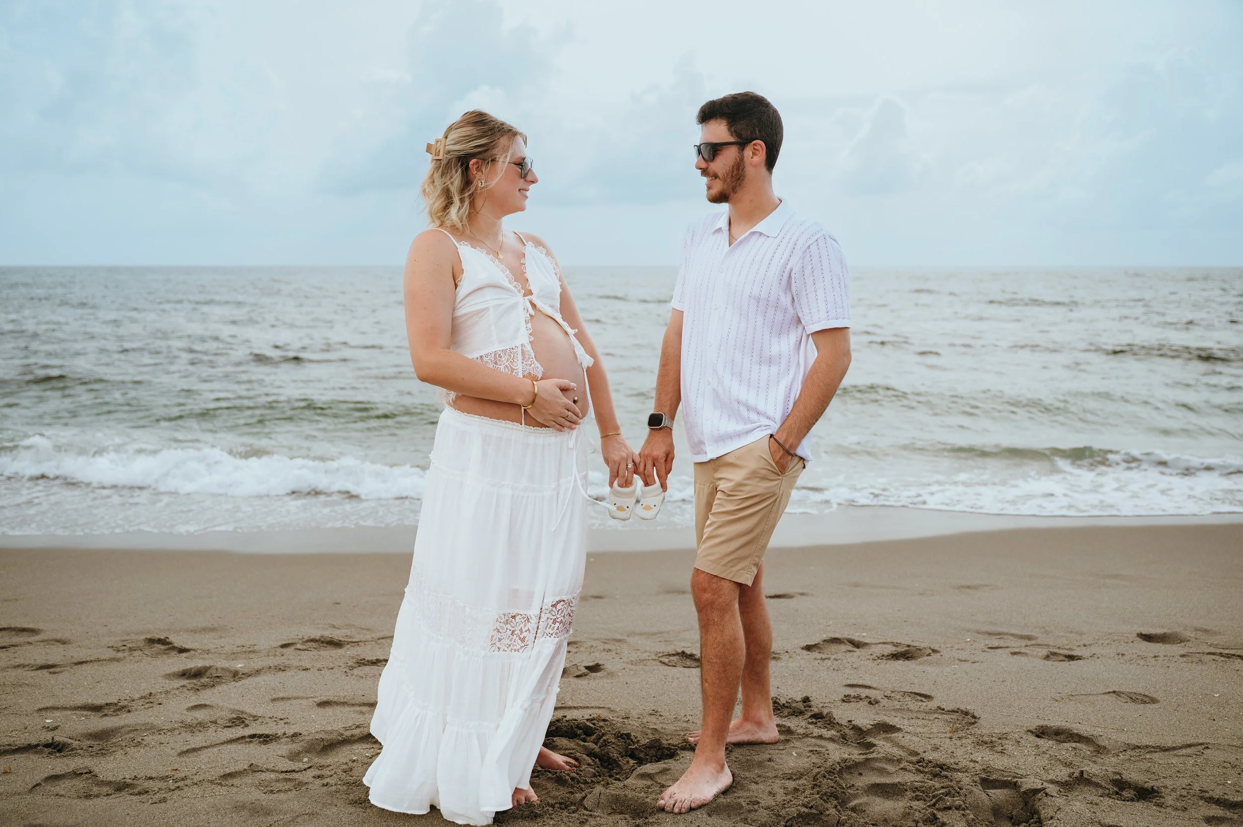 Expecting couple holding hands and tiny baby shoes during their beach maternity session at Blowing Rocks Preserve in Hobe Sound, Florida.