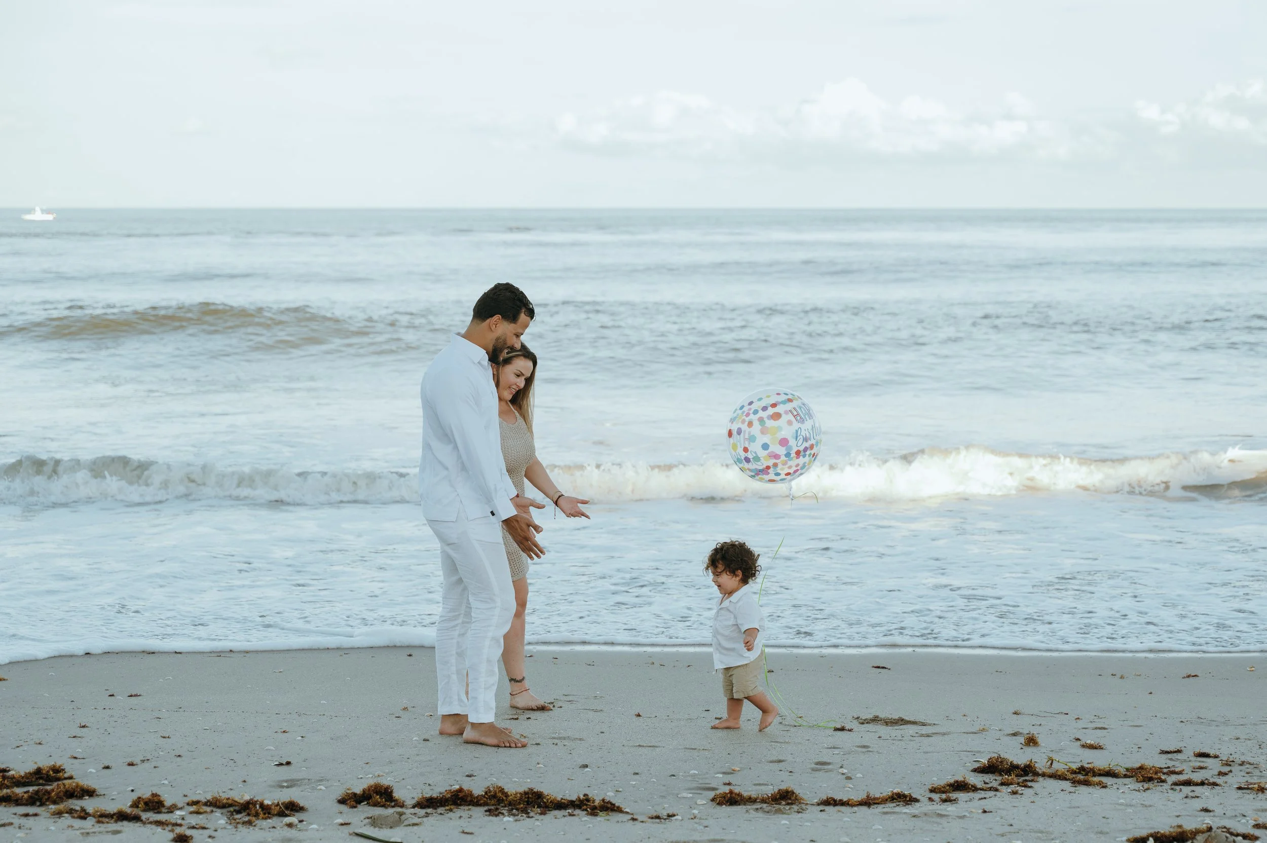 Parents watching their toddler run beside a birthday balloon during a family beach session at Boynton Inlet.