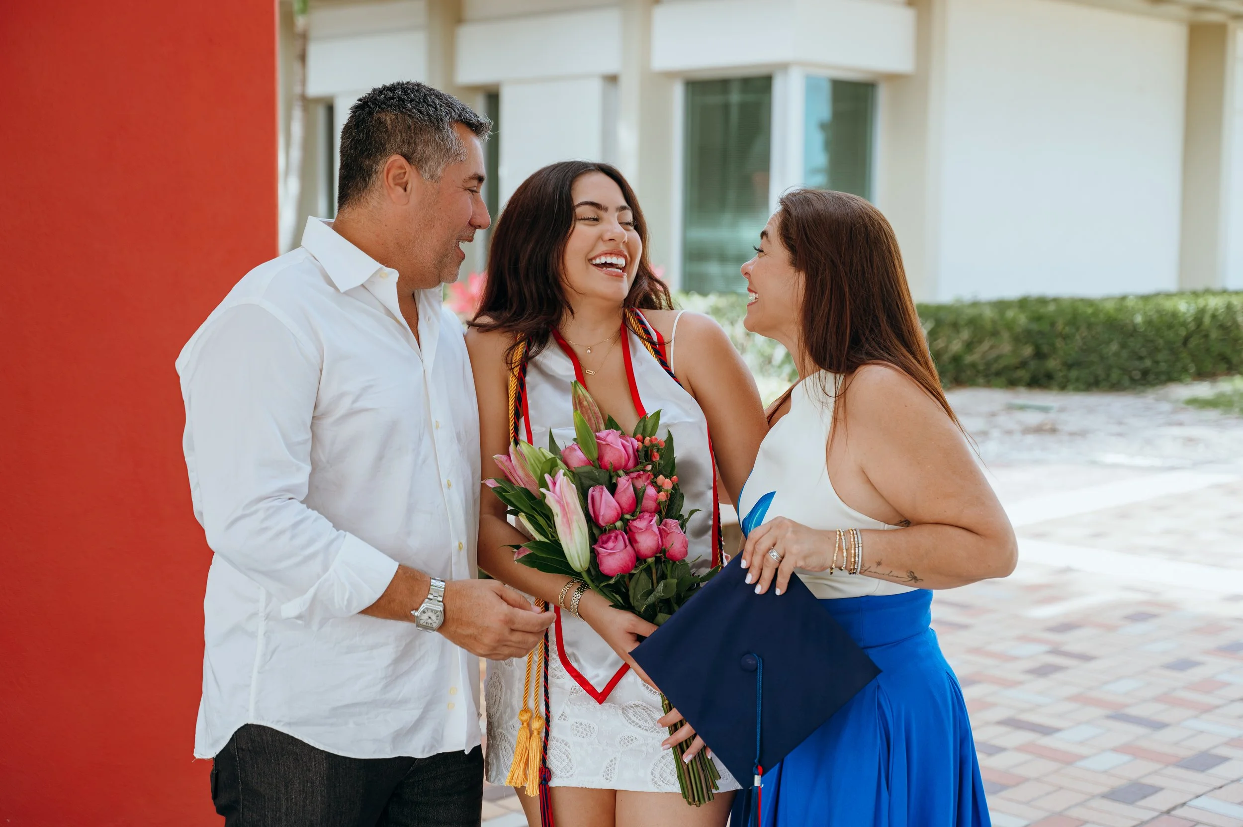 Florida Atlantic University graduate laughing with her parents while holding flowers and her graduation cap during her session in Boca Raton, Florida.