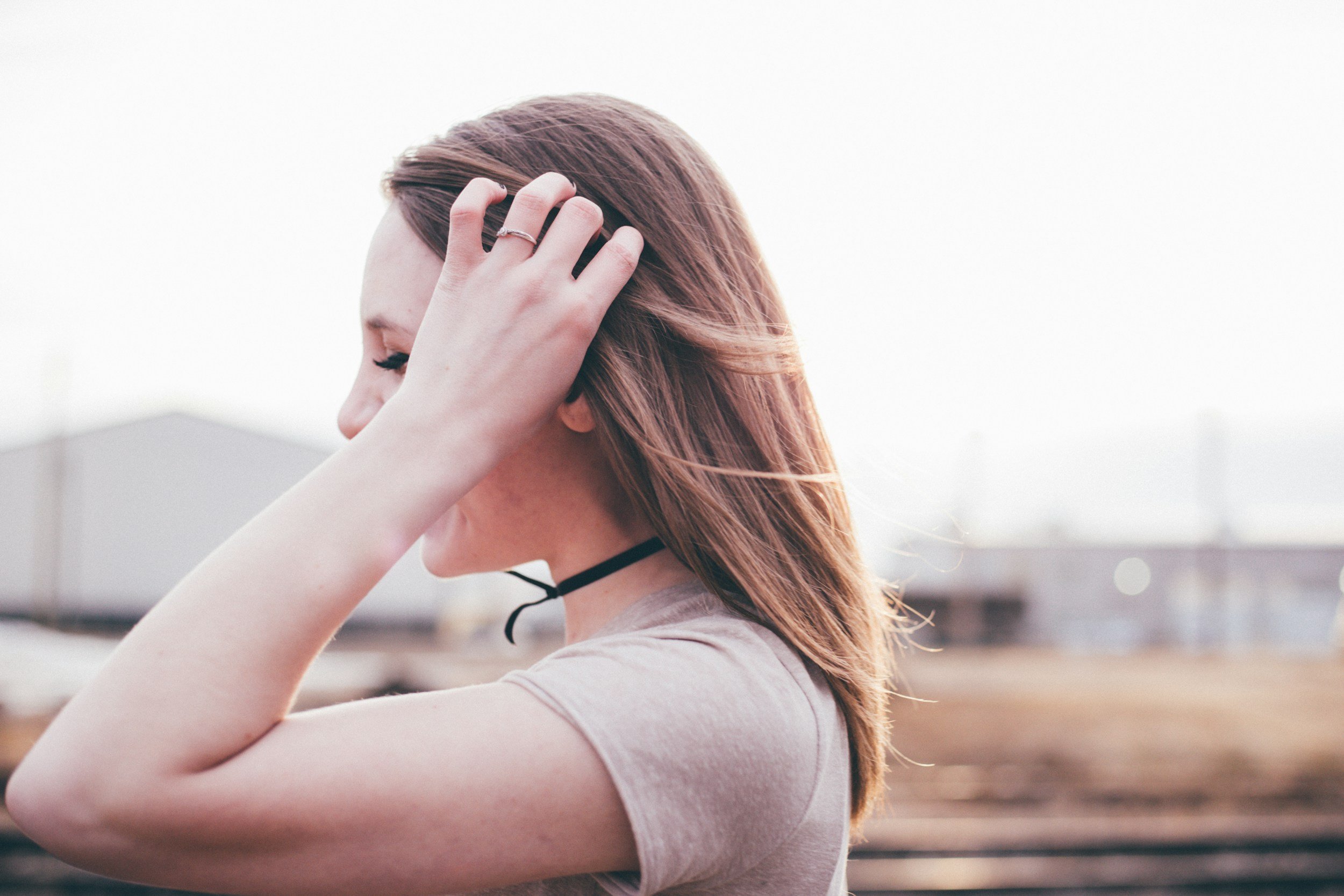 Side profile of a young woman with medium-length reddish-brown hair, wearing a beige t-shirt and a thin black choker, holding her head with one hand while outdoors with blurred background.