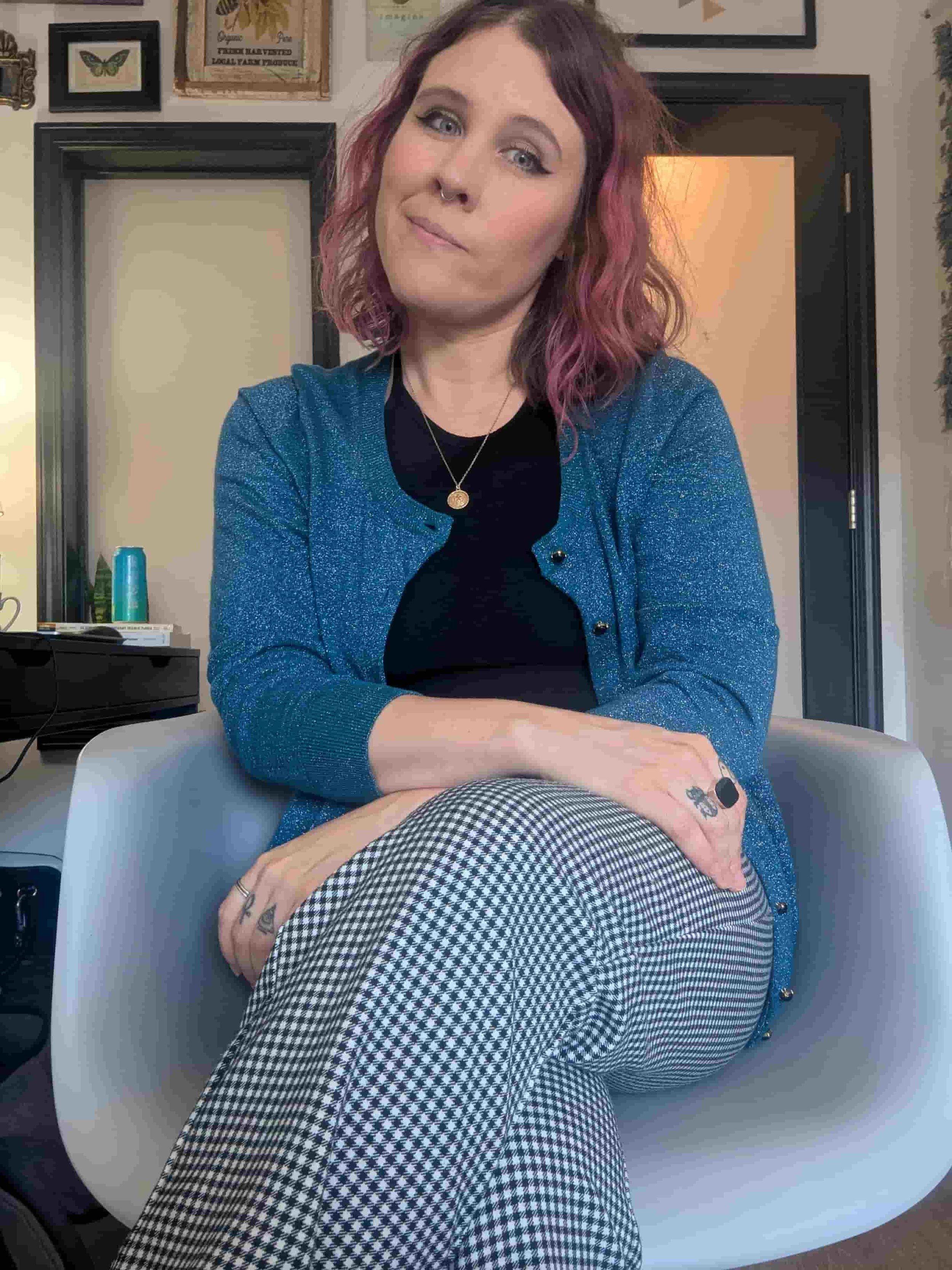 A woman with wavy pink and brown hair sitting on a white chair in a room with framed pictures on the wall. She is wearing a blue cardigan, black top, black and white checkered pants, layered rings and necklace, and has a septum piercing.