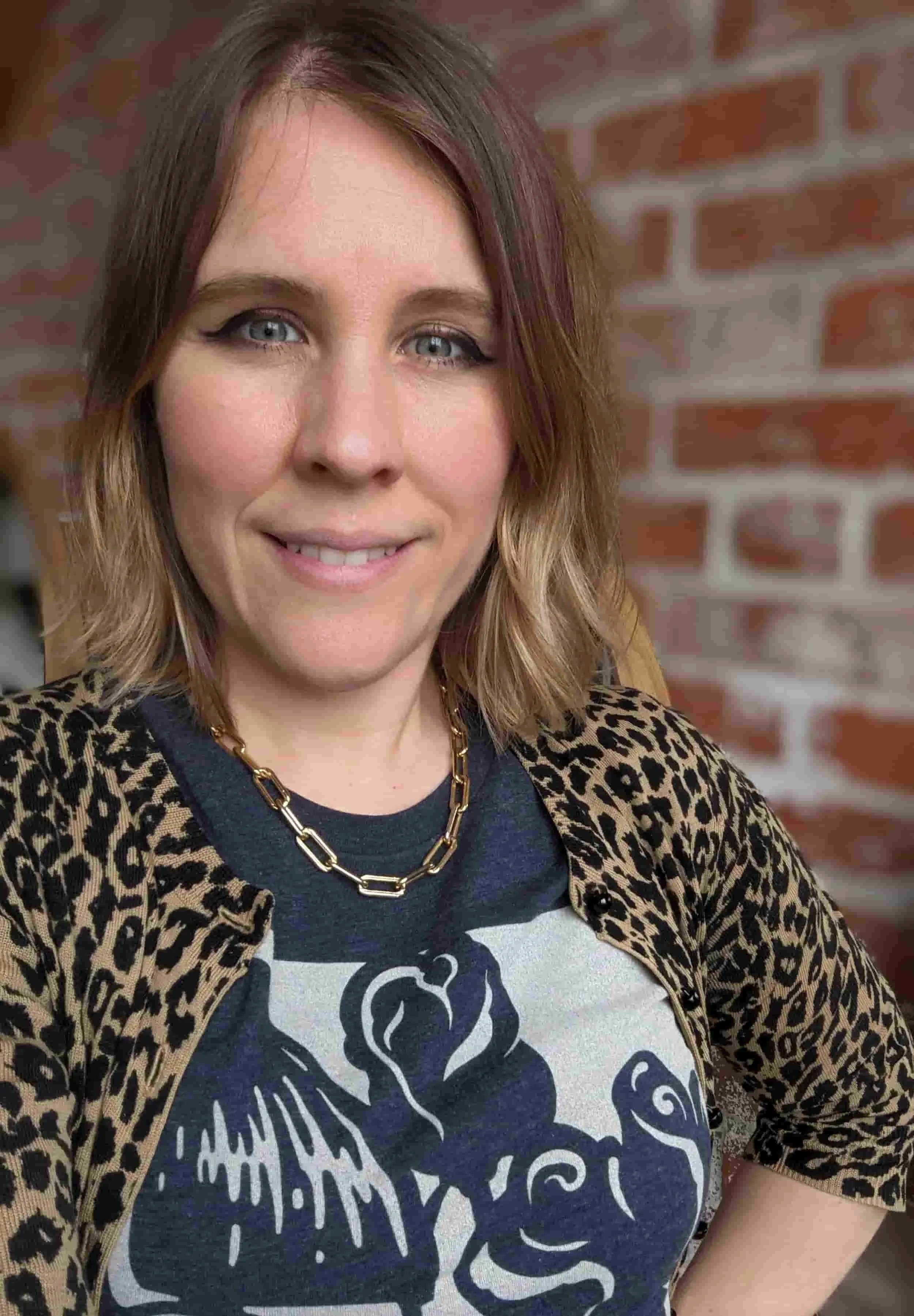 Trauma therapist Christin Trott. A woman with shoulder-length wavy hair, wearing a leopard-print top and a gold chain necklace, smiling in front of a brick wall.