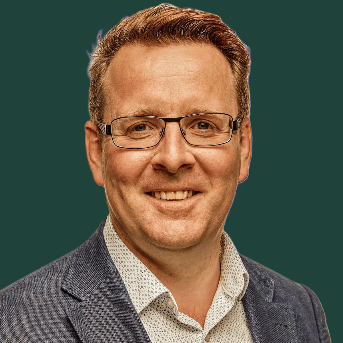 Close-up of a Edwin Eve wearing glasses, a patterned shirt, and a gray blazer, smiling against a plain dark green background.