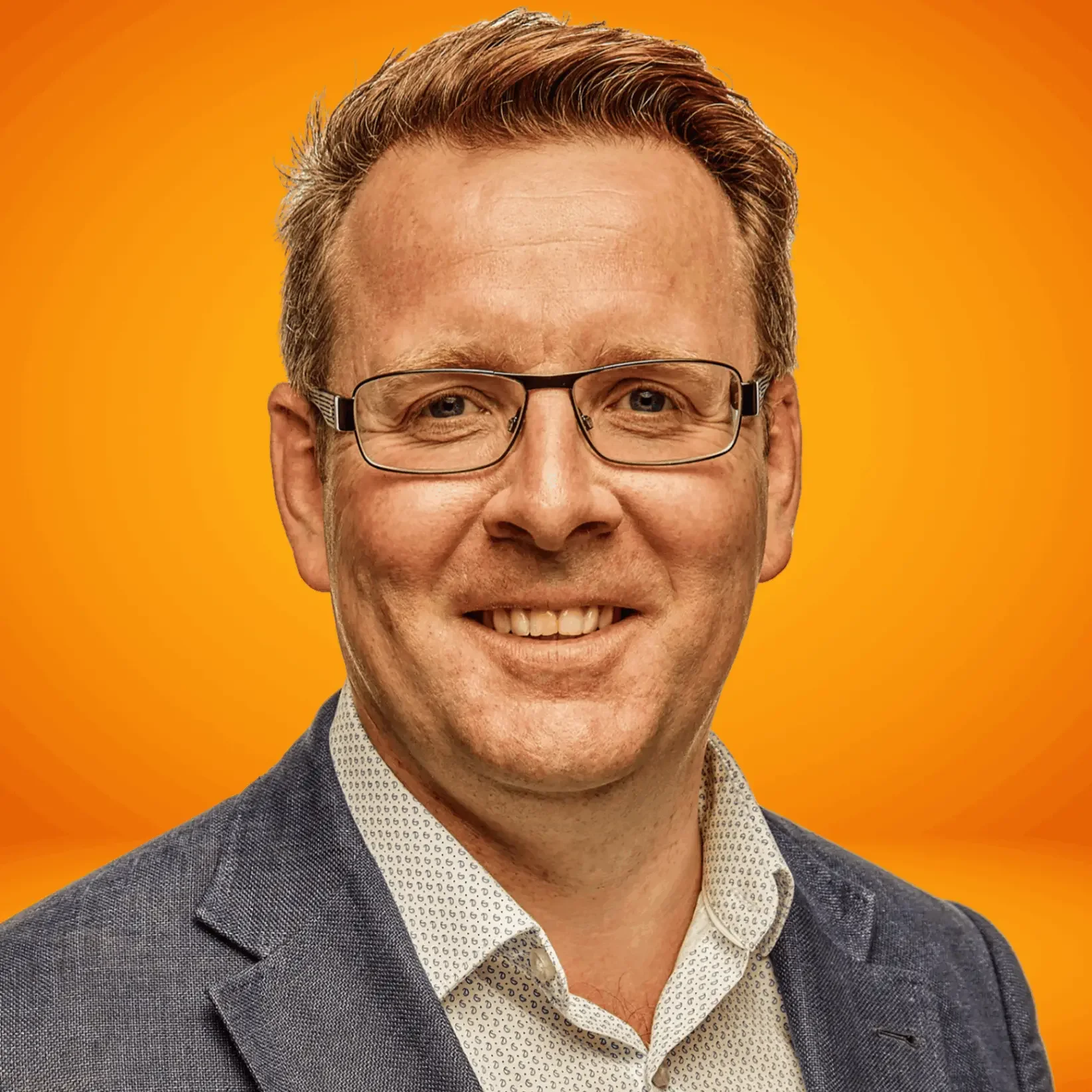 Edwin Eve, founder and ICF accredited coach, Close-up portrait of a smiling man with glasses, wearing a suit jacket and a patterned shirt, against an orange background.