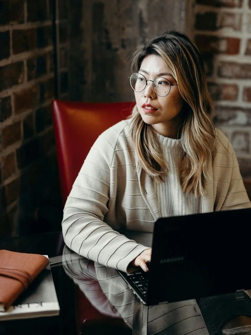 A woman pauses while working on her laptop, composed and capable but with her mind somewhere else.