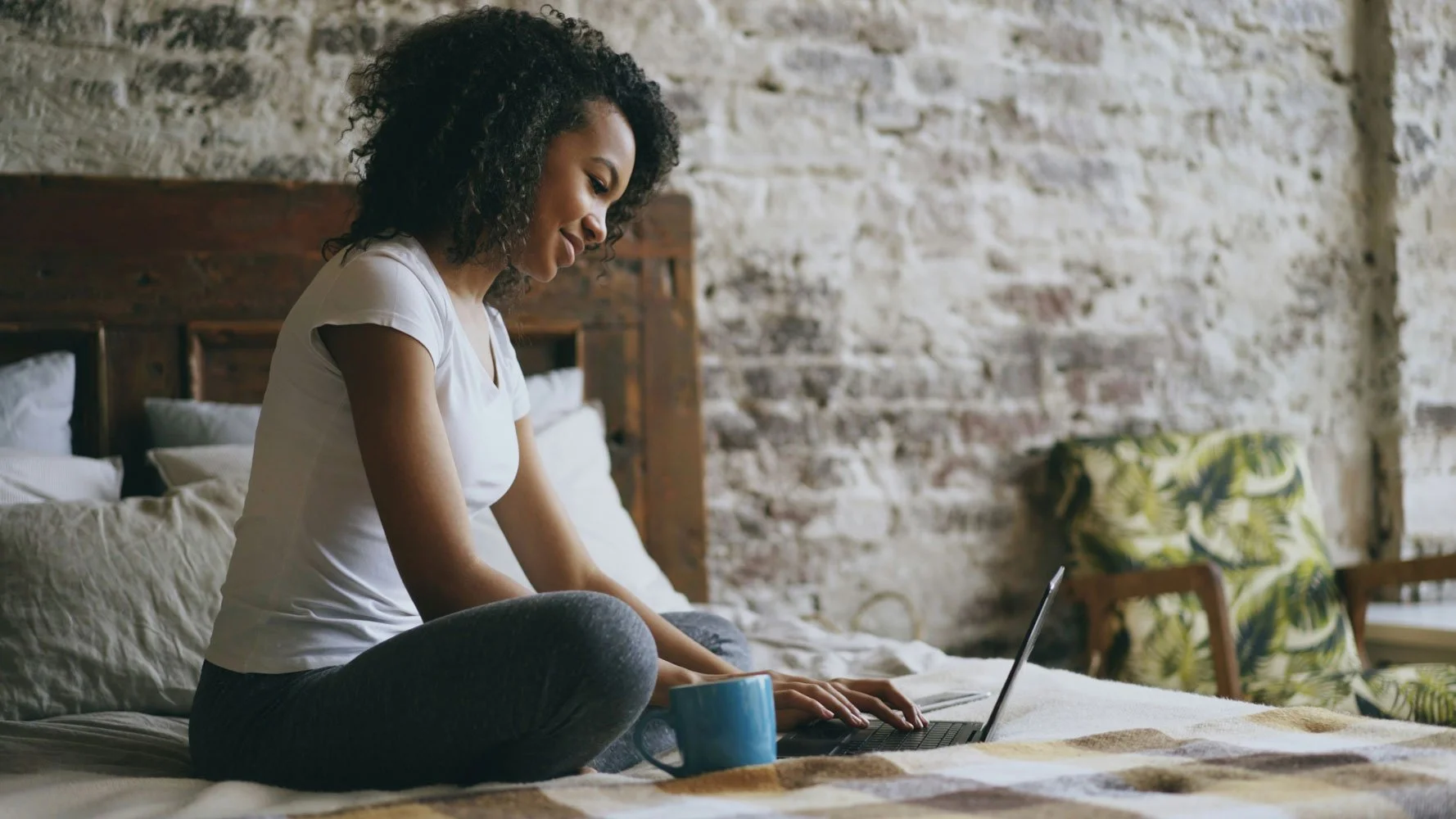 Woman sitting cross-legged on her bed with a laptop and a mug, in a relaxed home setting — representing the flexibility of attending online therapy from wherever you are