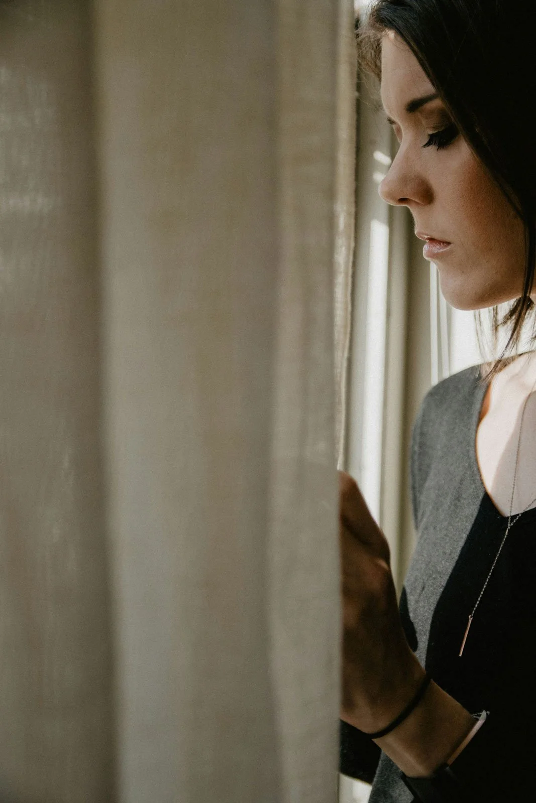 A woman stands close to the curtain at her window, glancing out, safe at home but with her guard still up.