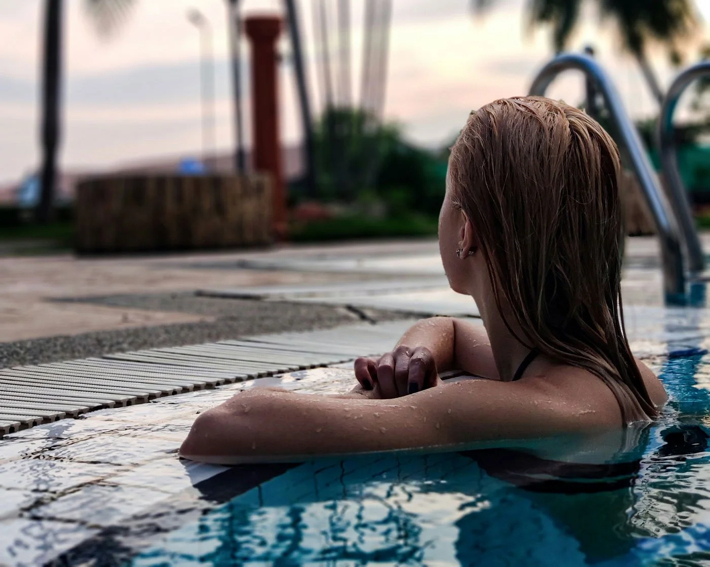 Woman resting at the edge of a pool at dusk, looking out into the distance with a quiet, faraway expression — capturing the feeling of grief in the middle of a season that expects you to be okay