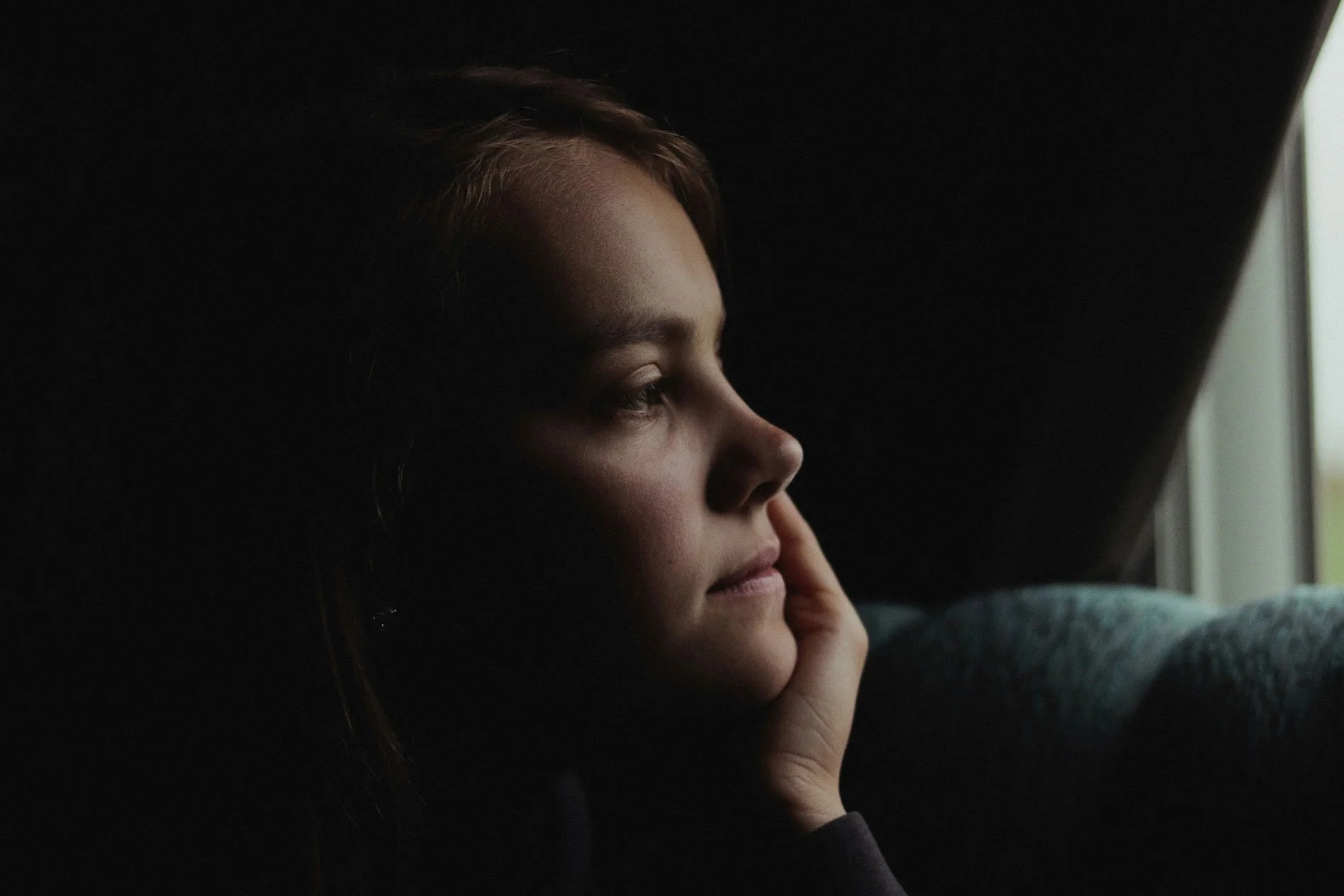 A woman rests her chin on her hand and gazes out a window in a dark room, sitting with grief that has surfaced months after her loss.