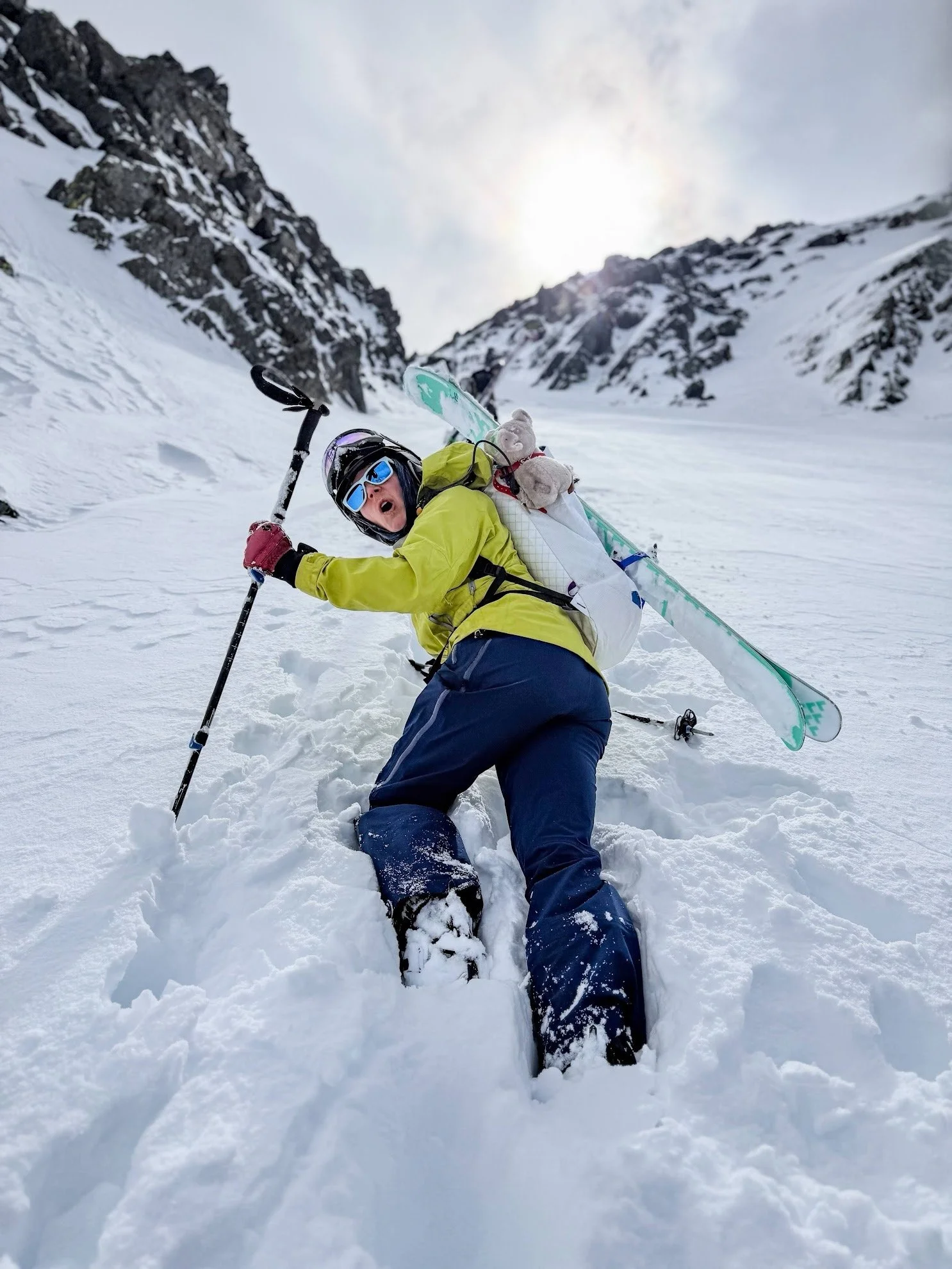 Person lying on snow-covered mountain slope holding skis and ski poles, wearing a yellow jacket, blue pants, helmet, and goggles, with rocky mountain cliffs in the background under a cloudy sky.