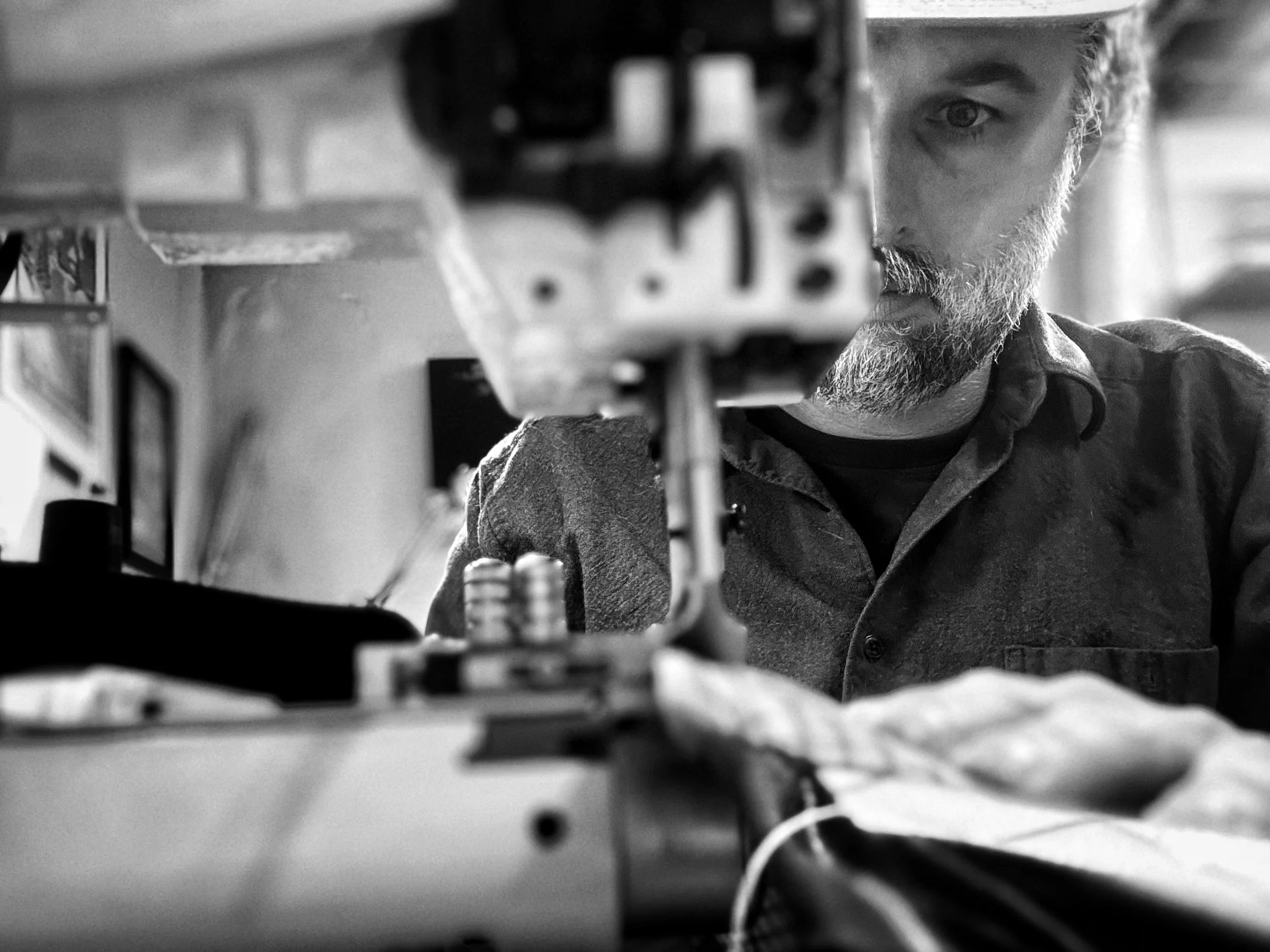 Black and white photo of a man with a beard looking through machinery or equipment, partially obscured and in focus.