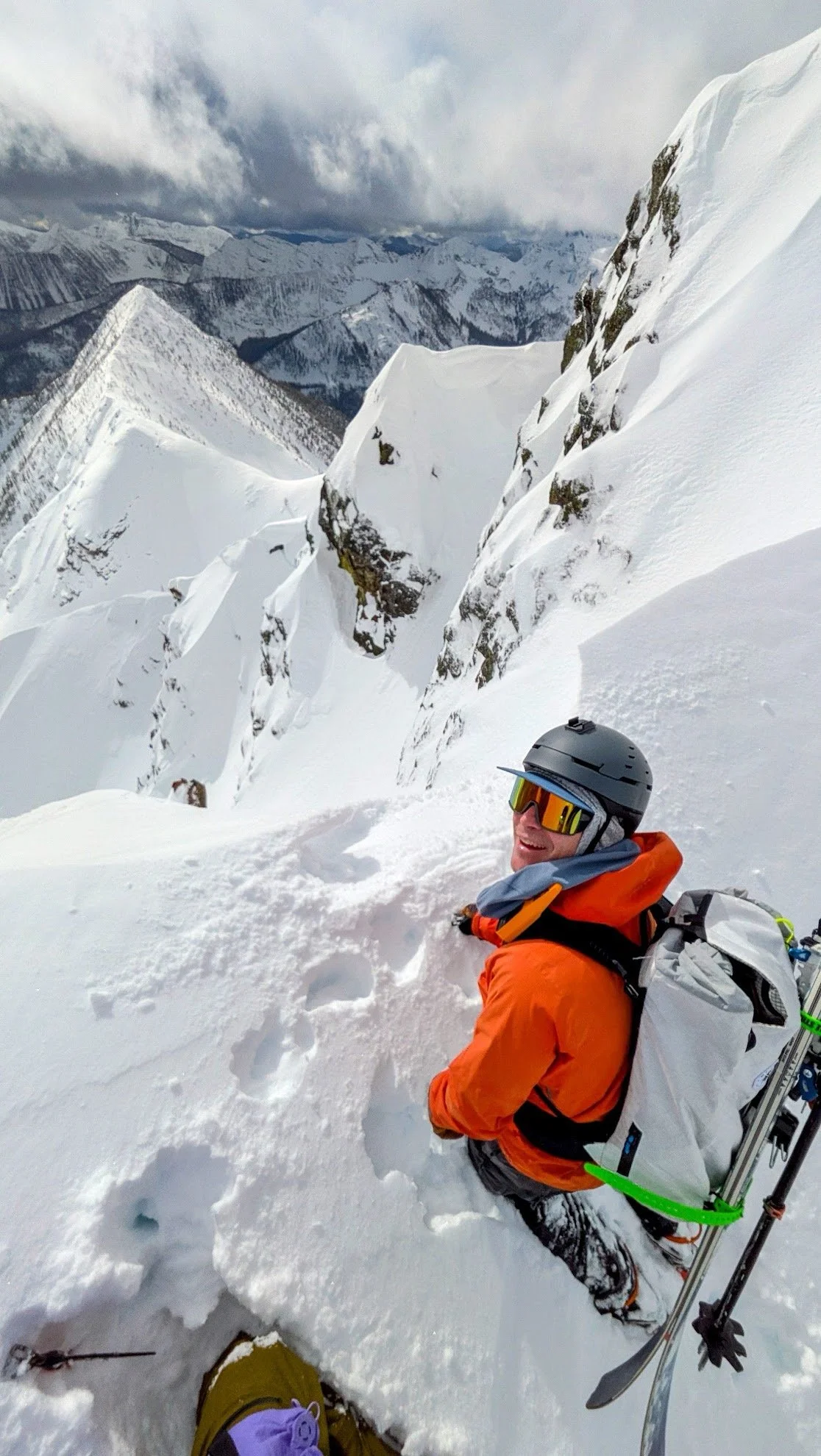 A person in orange winter gear, wearing a helmet and sunglasses, sitting on snow on a mountain slope, with a snowy mountain range in the background.