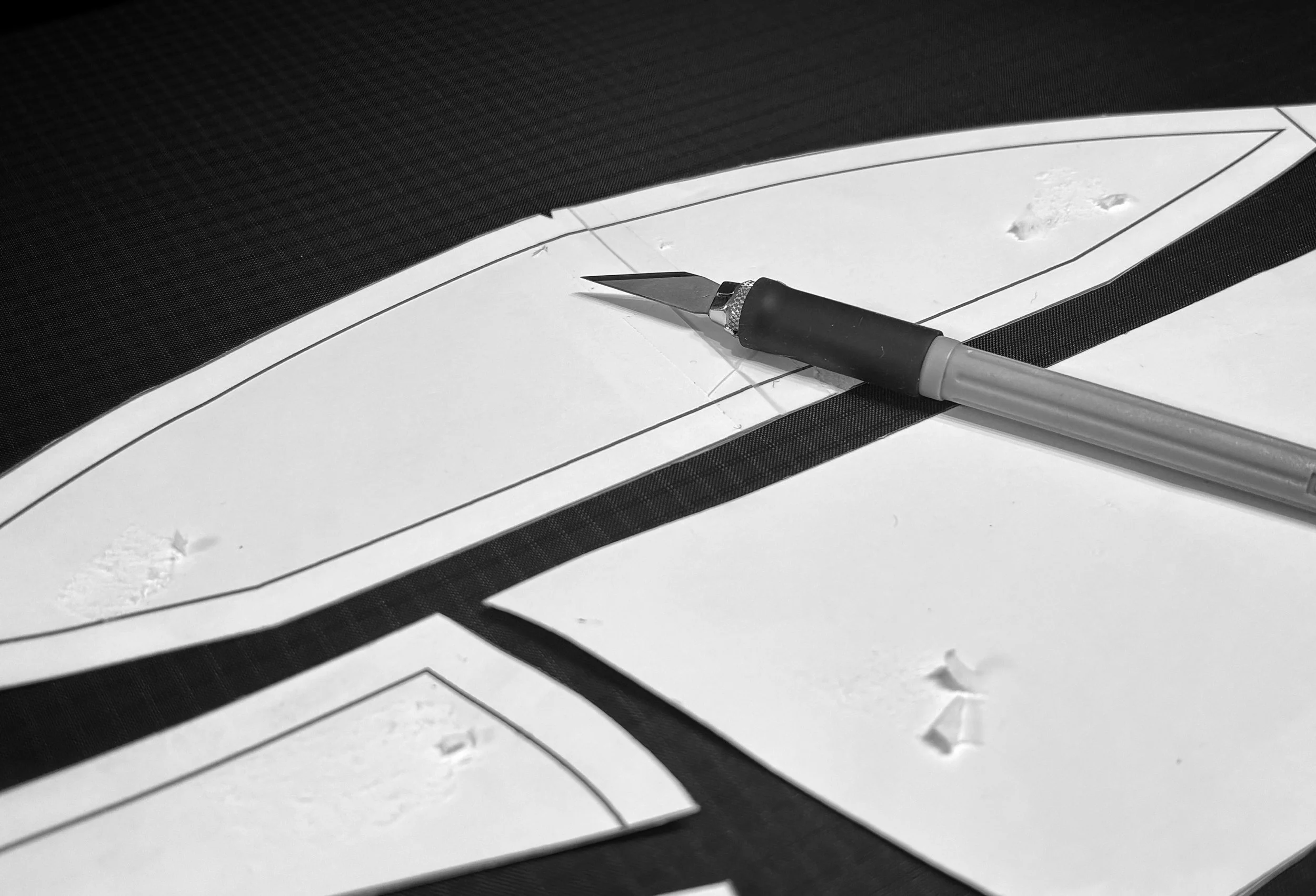 A black and white photograph of a utility knife placed on a craft paper with printed outlines for cutting or folding.