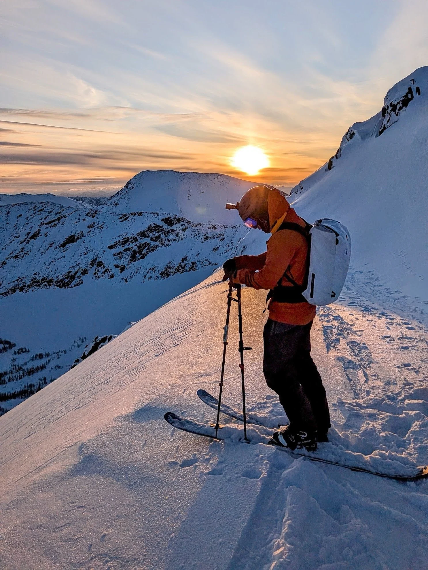 A person dressed in orange winter gear and a helmet standing on snowy mountain slope with a sunset in the background.