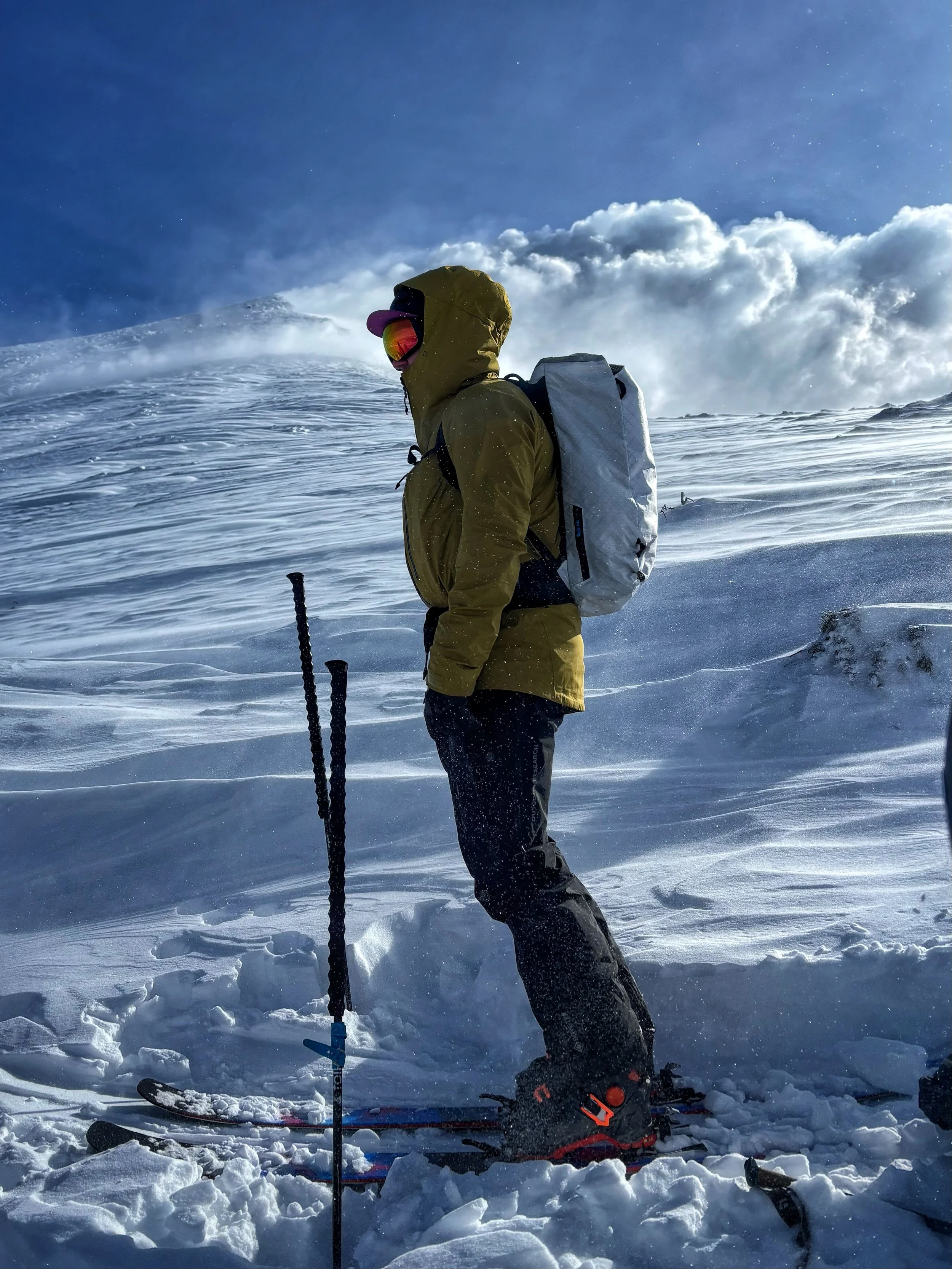 A person standing on snow-covered ground in winter gear, including a brown hooded jacket, snow goggles, and black snow boots. They are holding ski poles and have skis attached to their boots, with a backpack on their back. The sky is partly cloudy with blue sky and white clouds, and snow is being blown around.