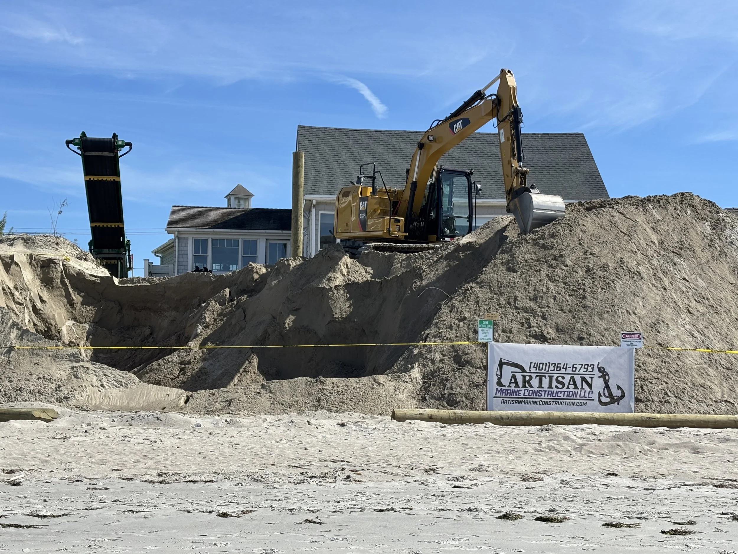 Construction site with a yellow excavator on top of a dirt mound, a large strip of sand in the foreground, and a house with a gray roof and a small tower in the background under a blue sky.