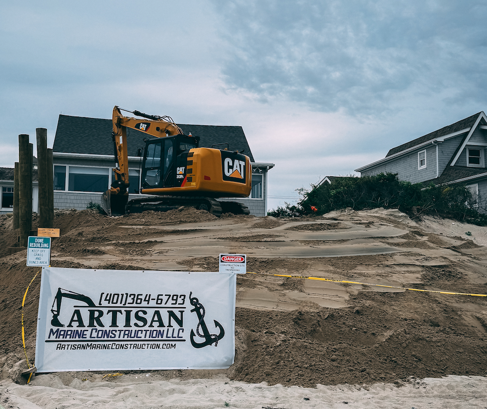 Construction site on a sandy hill with a yellow Cat excavator and signs from Artisan Marine Construction LLC, with houses in the background and cloudy sky.