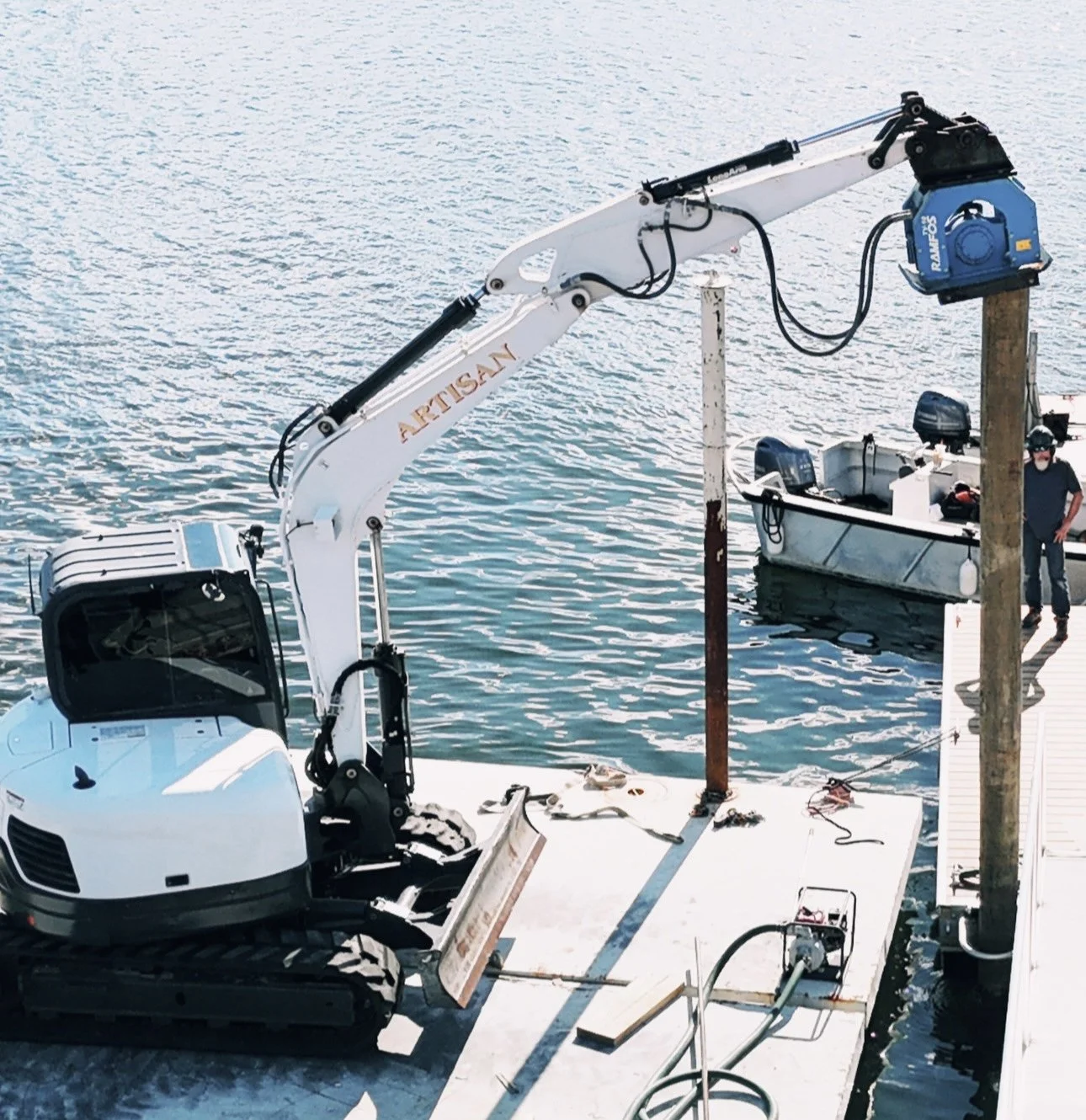 A small robotic excavator on a dock using a crane arm to lift a wooden post out of the water.