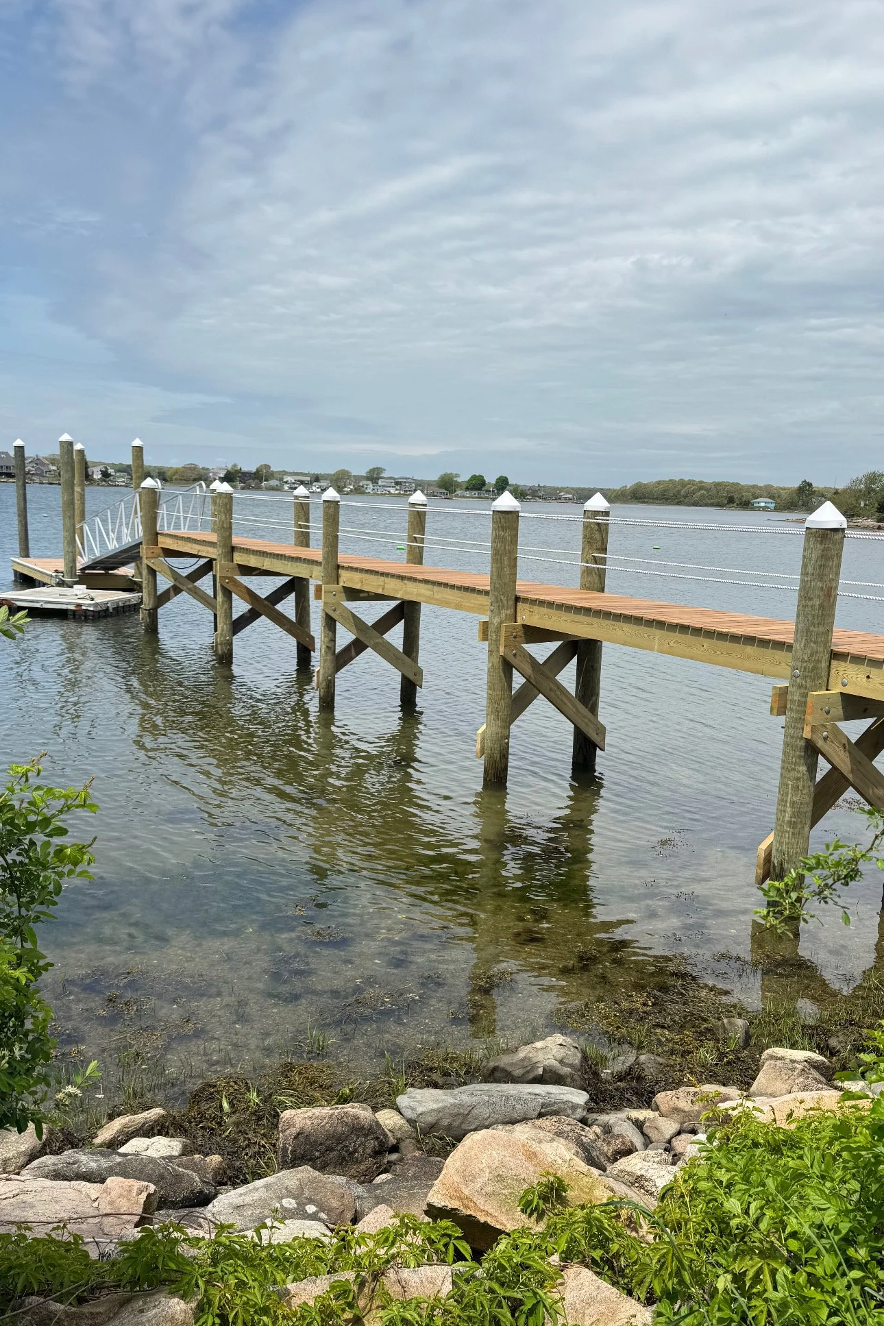 A wooden dock extending into a calm body of water, with rocks and green plants along the shoreline in the foreground, and a partly cloudy sky overhead.