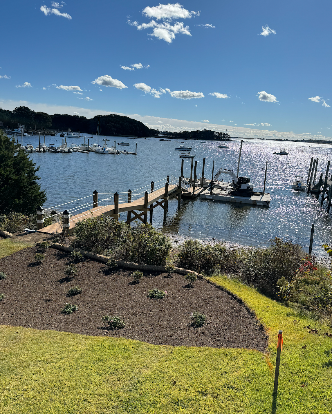 View of a marina with boats docked on a sunny day, a wooden pier extending into the water, surrounded by landscaped garden with newly planted bushes, and a clear sky with scattered clouds.