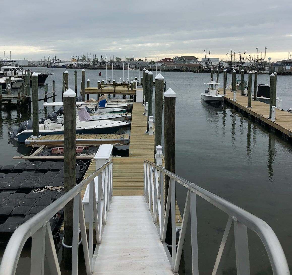 A marina with boats docked at wooden piers on a cloudy day, viewed from a white metal gangway leading down to the docks.