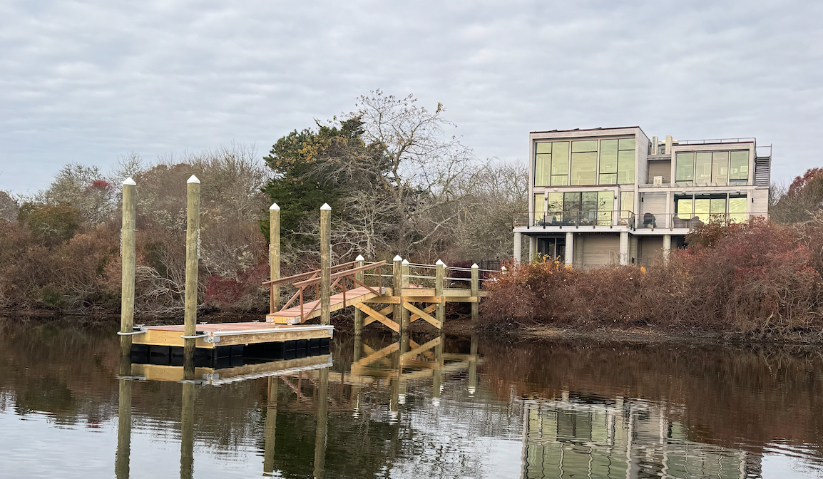 Modern house with large windows on the waterfront, a small wooden dock with boat slips, and a cloudy sky.