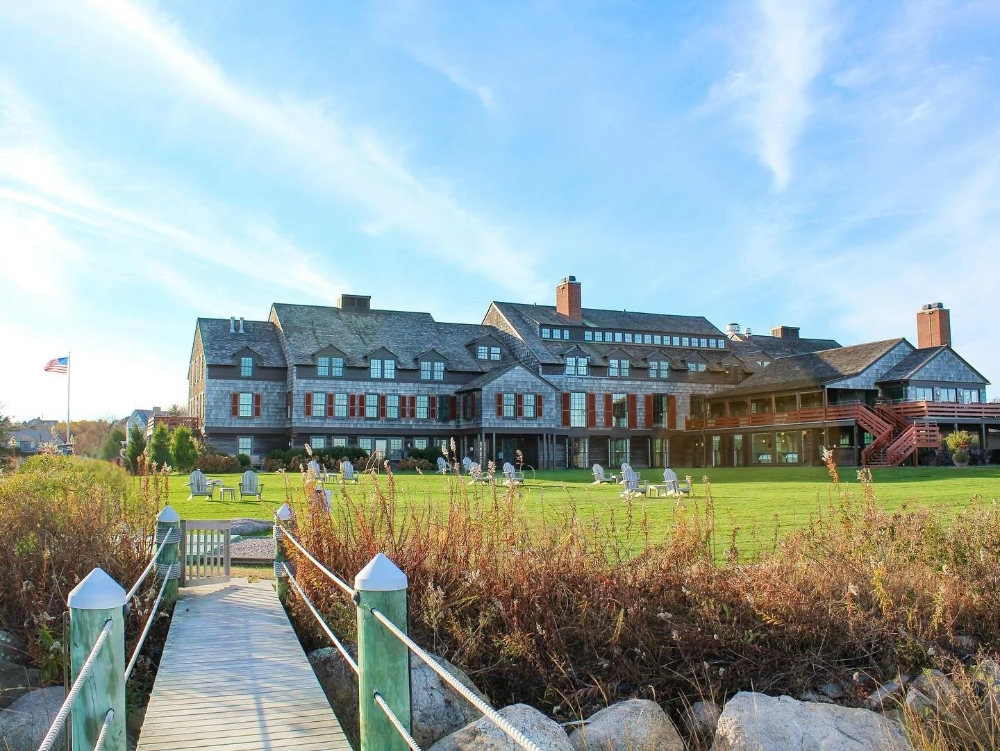 Large gray multi-story building with red shutters, surrounded by a green lawn, Adirondack chairs, and a wooden walkway leading to a landscaped area, under a partly cloudy sky.
