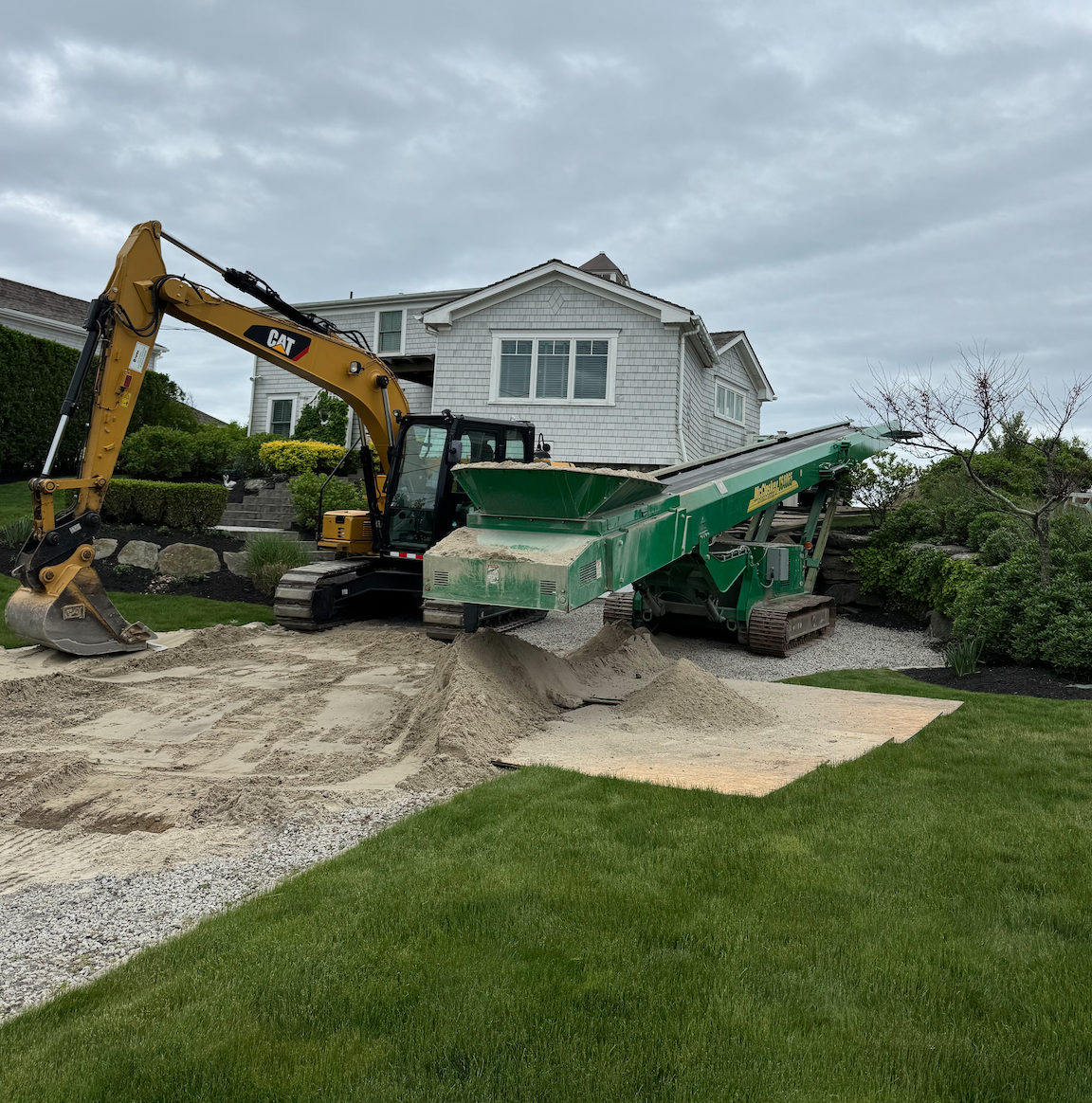 Construction equipment, including a yellow excavator and a green soil screener, working on a landscaped yard with a mound of sand, a house, and cloudy sky.