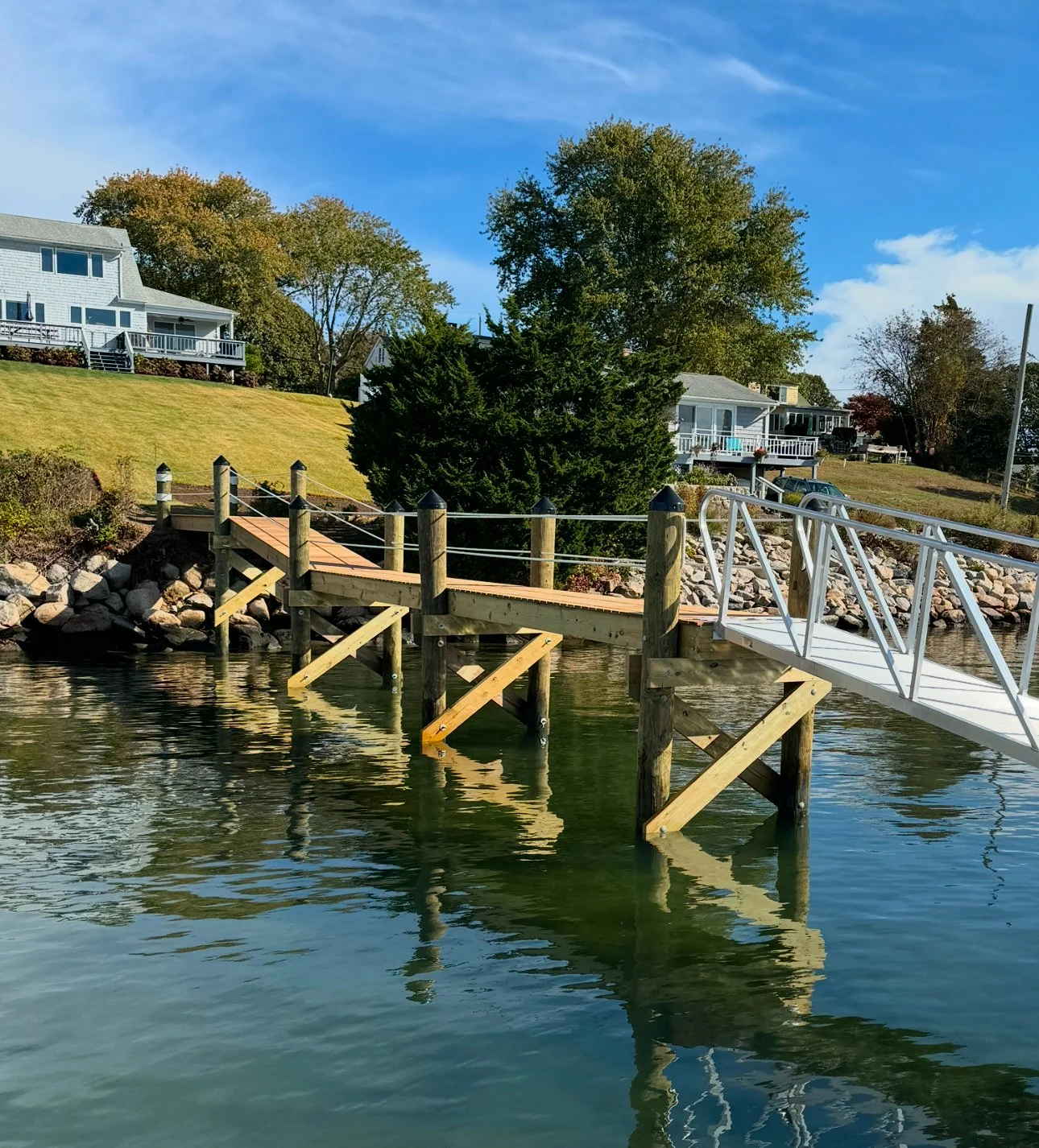 A wooden dock extending into a body of water with houses and trees in the background under a blue sky.
