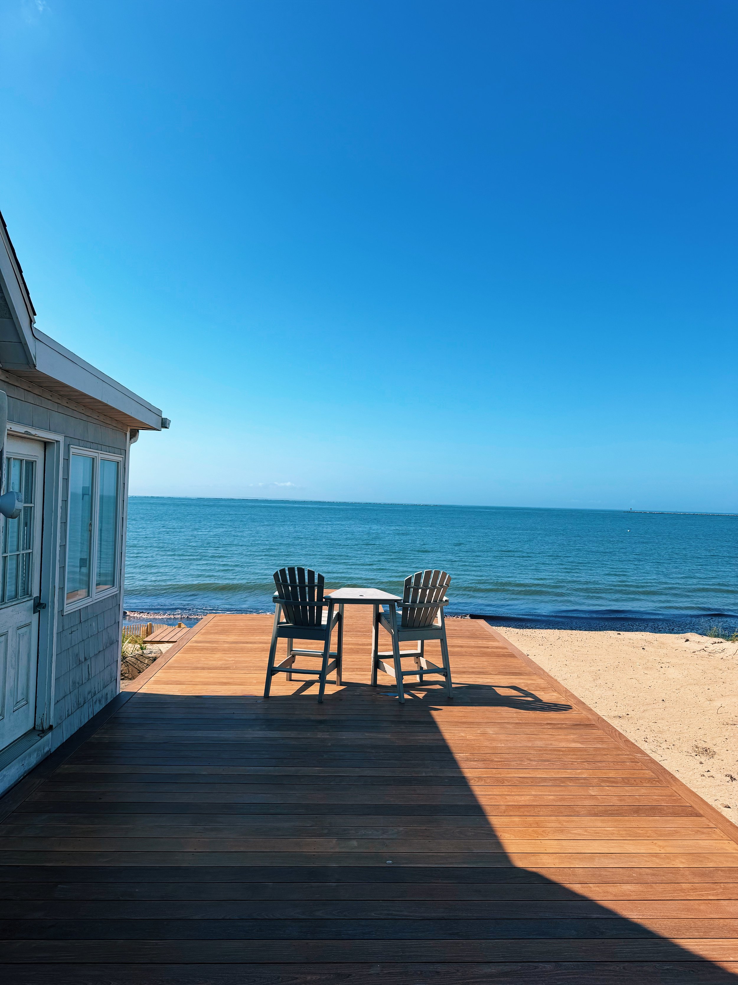 Empty wooden deck with two chairs and a small table overlooking the ocean under a clear blue sky, with part of a beach house on the left.