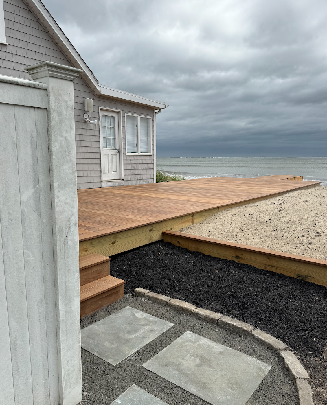 A newly built wooden deck attached to a house by the beach, with steps leading up from a concrete pathway, over black gravel, overlooking the ocean under cloudy sky.