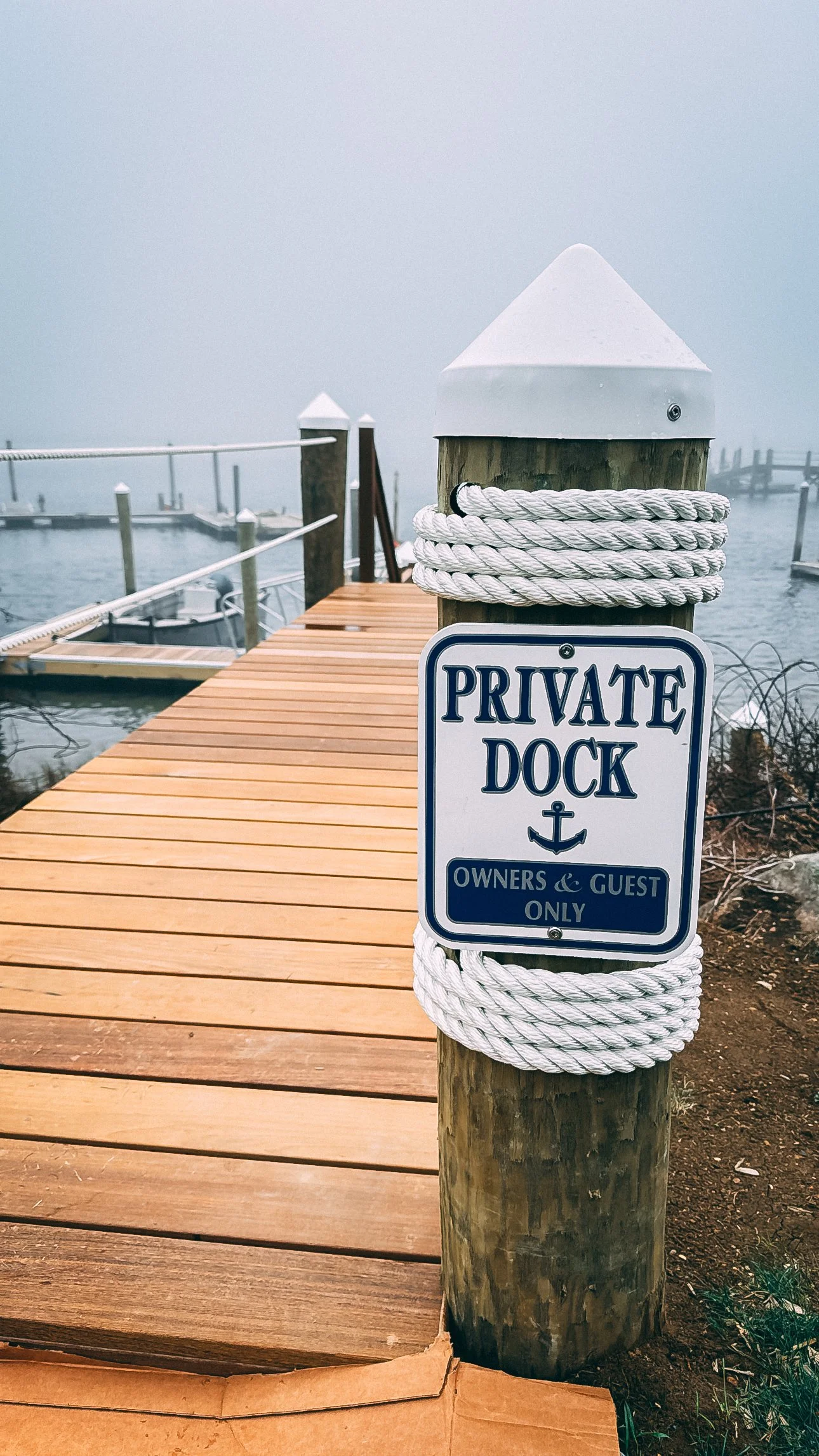 A wooden dock with a sign reading 'Private Dock Owners & Guest Only' wrapped around a post with ropes. The dock extends into a foggy marina with boats and piers.