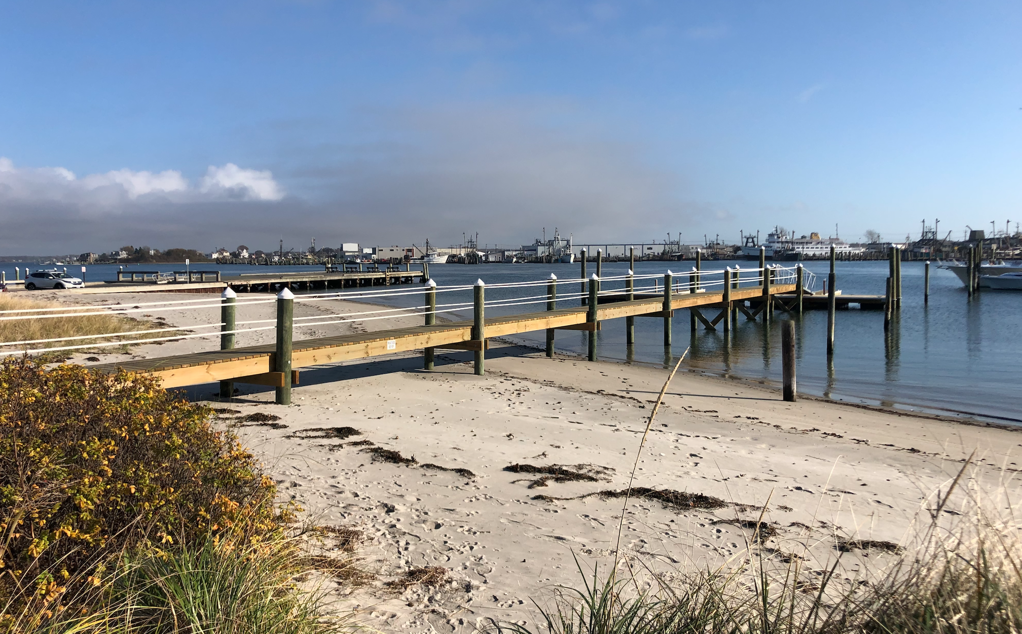 A sandy beach with a wooden pier extending into calm water, boats docked in the harbor, and a sky with scattered clouds.