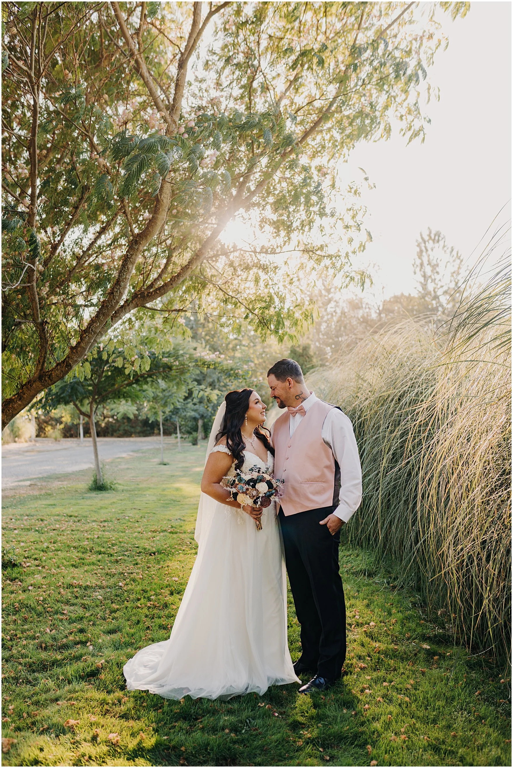 A bride and groom standing together outdoors in a park-like setting, facing each other and smiling, with greenery and tall grasses around them and sunlight filtering through trees overhead. Green Villa Barn, Salem Oregon