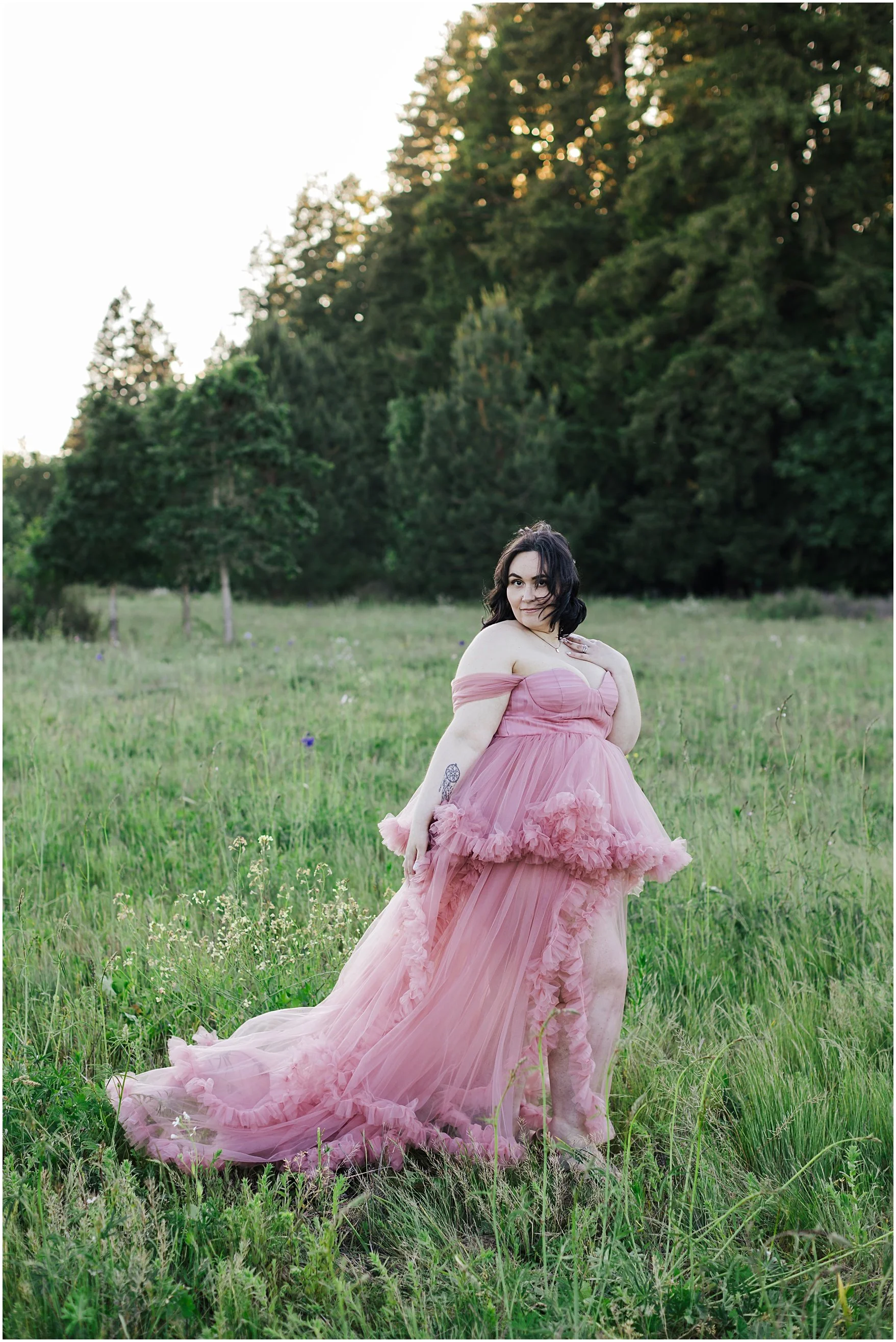 Woman in a flowing pink gown standing in a grassy field with trees in the background during sunset.
