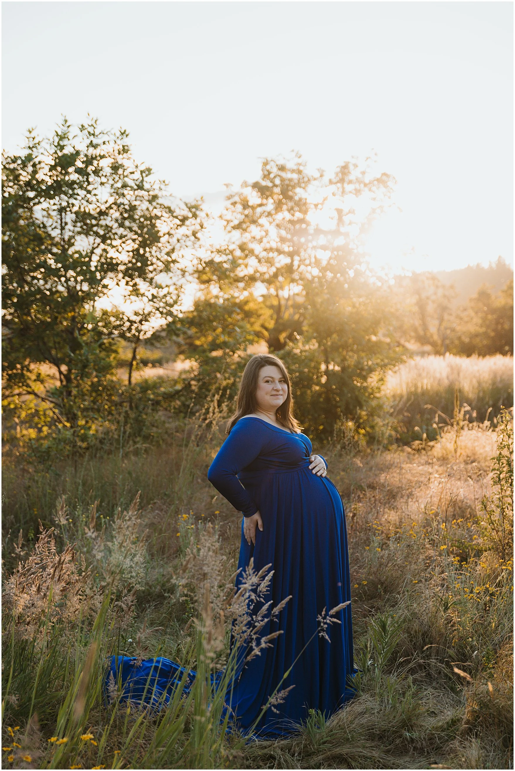 A pregnant woman in a long blue dress stands in a field of tall grass and wildflowers, with trees and the setting sun in the background.