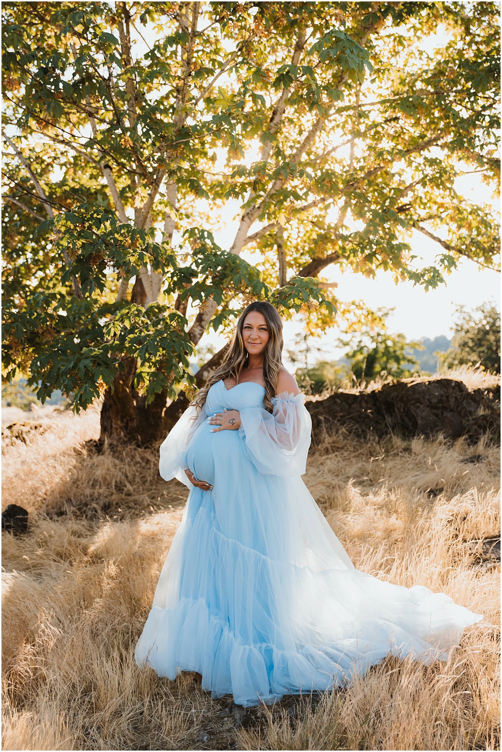 A woman in a light blue dress standing outdoors in a field of dry grass, with a large leafy tree behind her. The scene is lit by warm sunlight, creating a serene, scenic setting.