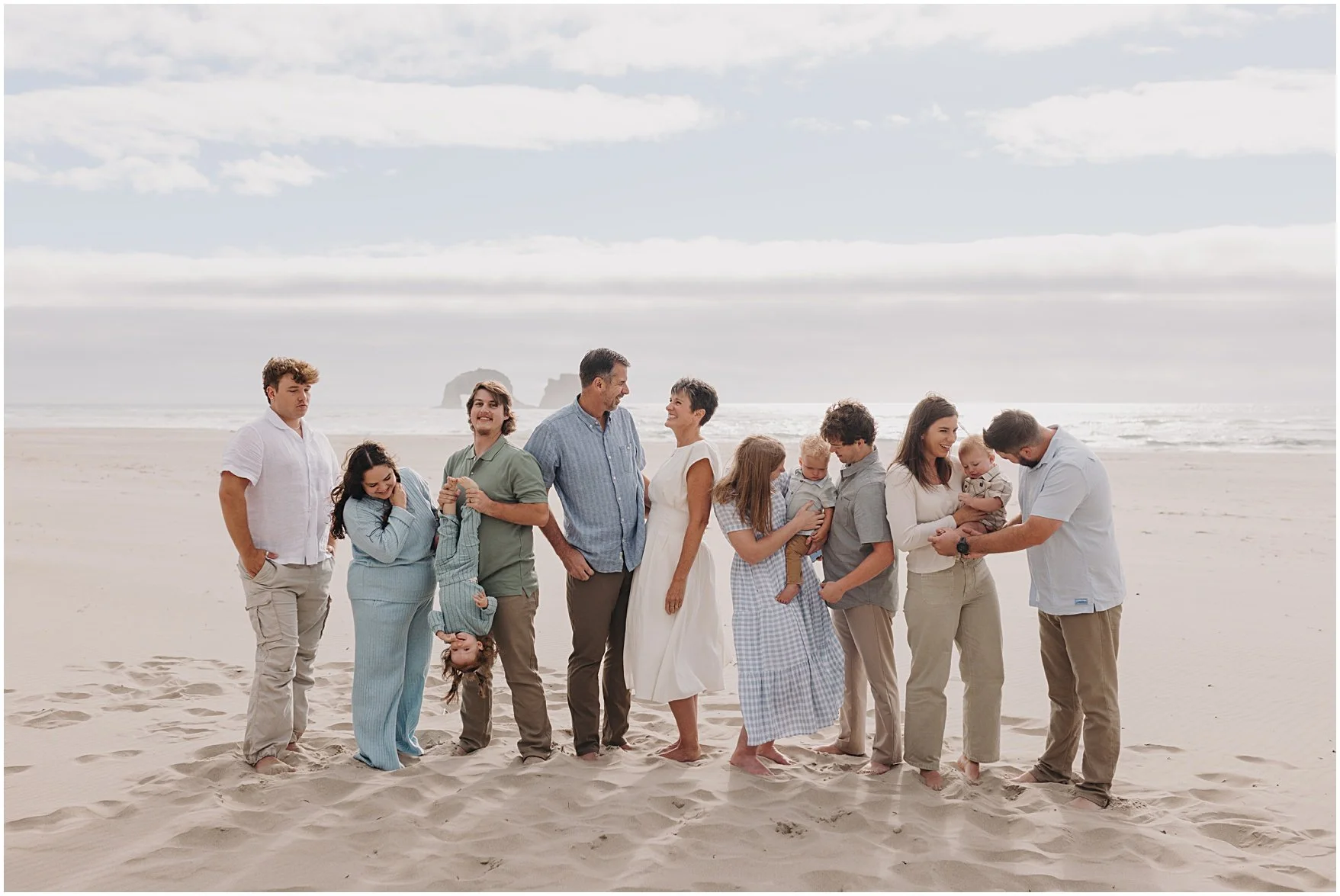 A diverse family gathering on the beach smiling and interacting with each other, some holding children, others standing and talking, under a cloudy sky with the ocean in the background. Rockaway Oregon